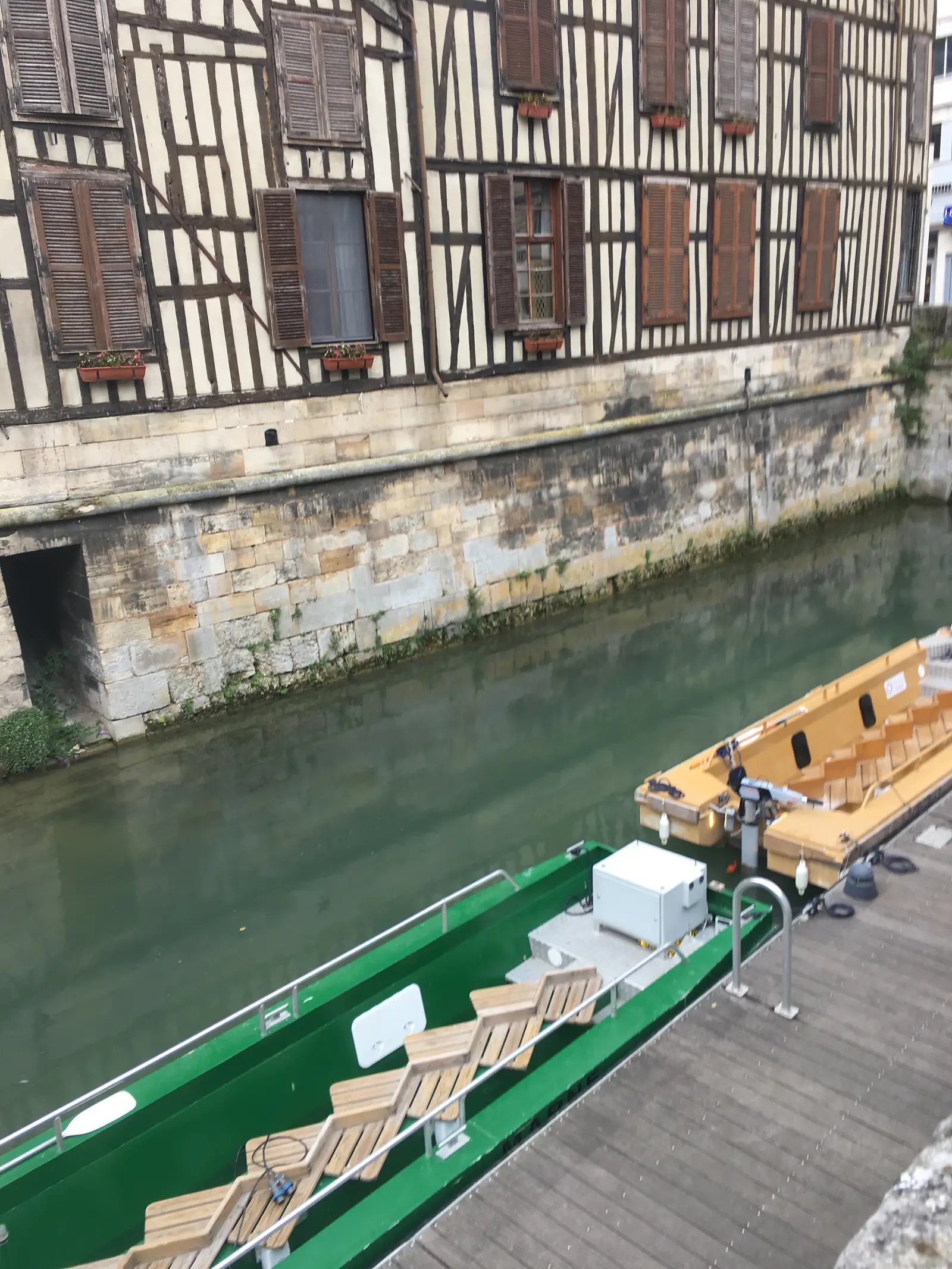 Looking down from a balcony onto a narrow town canal, a green tour boat with wooden deck chairs moored beside a half-timbered building.