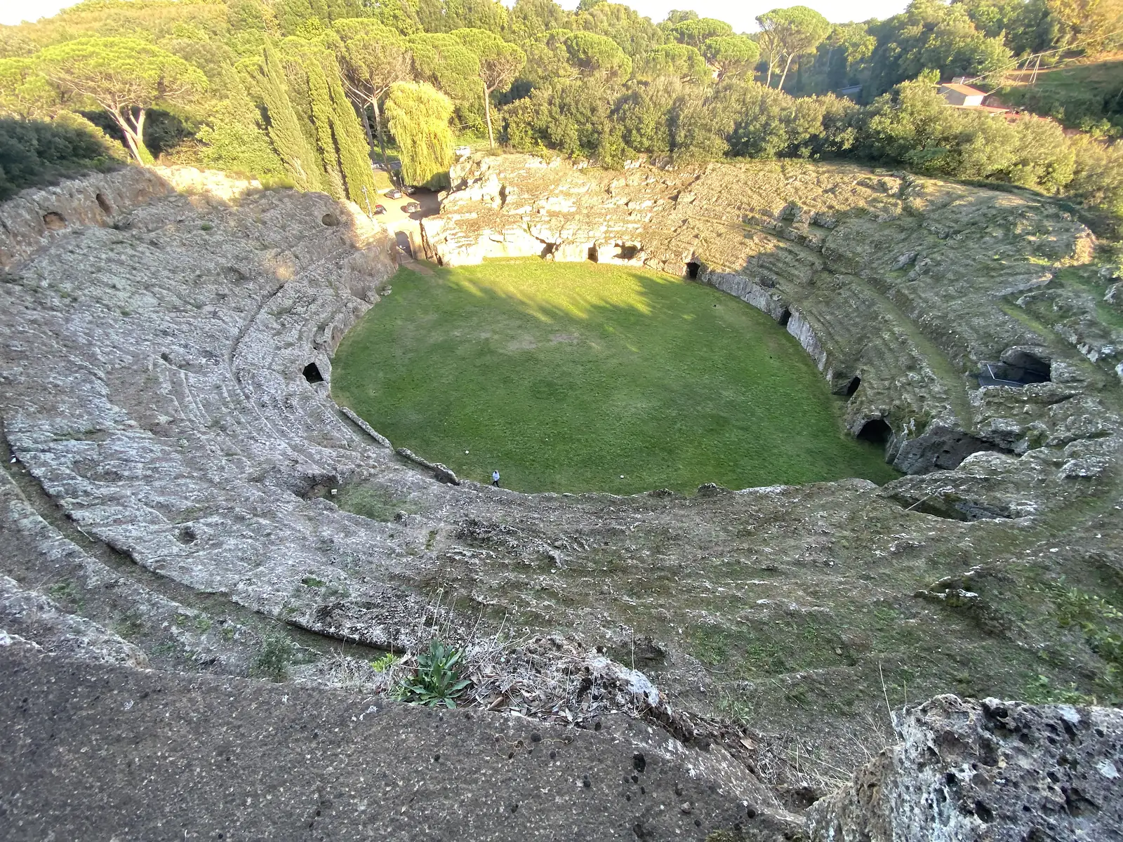 A high view down into a huge oval amphitheatre carved entirely from the rock, its tiered seating encircling a grassy arena where a single visitor stands tiny below, framed by pine trees.
