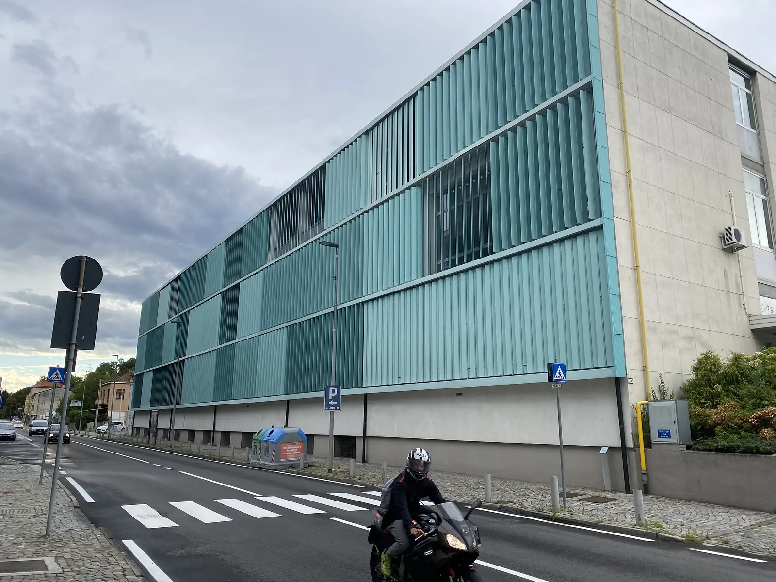 The turquoise, vertically louvred facade of a modern building on a street corner, a motorcyclist riding past in the foreground.