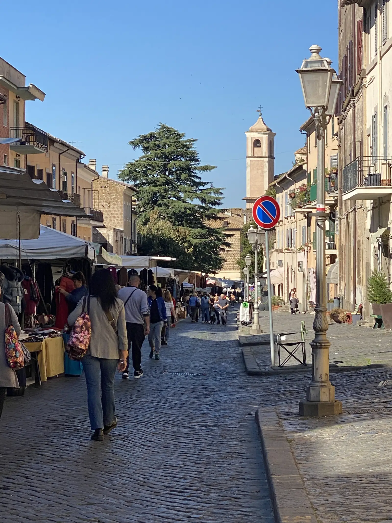 A busy Italian town square with a street market of stalls under white awnings, shoppers walking past on the paved piazza, a stone lamppost, a no-parking sign, and a bell tower against a clear sky.
