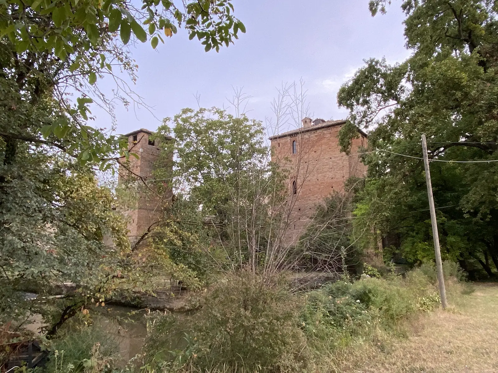 A brick medieval tower and an adjoining fortified building rise above a canal and dense trees, with a power line and pole in the foreground.