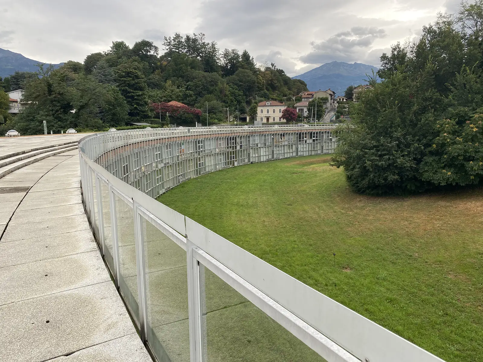 A curved, paved rooftop terrace with a tall aerial mast and domed skylights set into the lawn beside it, overlooking a low glazed building and wooded hills in the Aosta Valley.