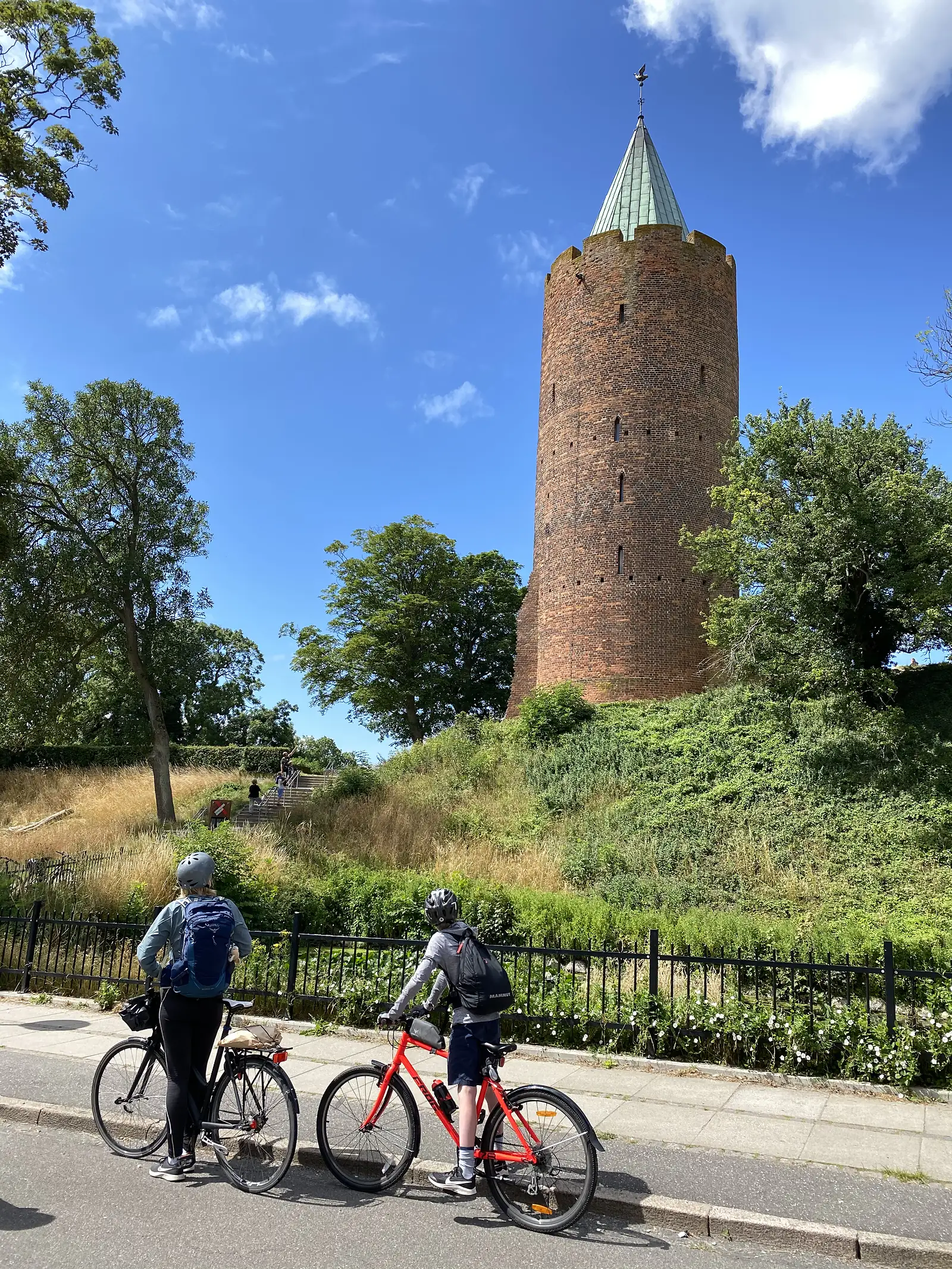 Two cyclists standing with their bikes on a pavement below a round red-brick medieval tower with a green copper spire on a grassy mound.