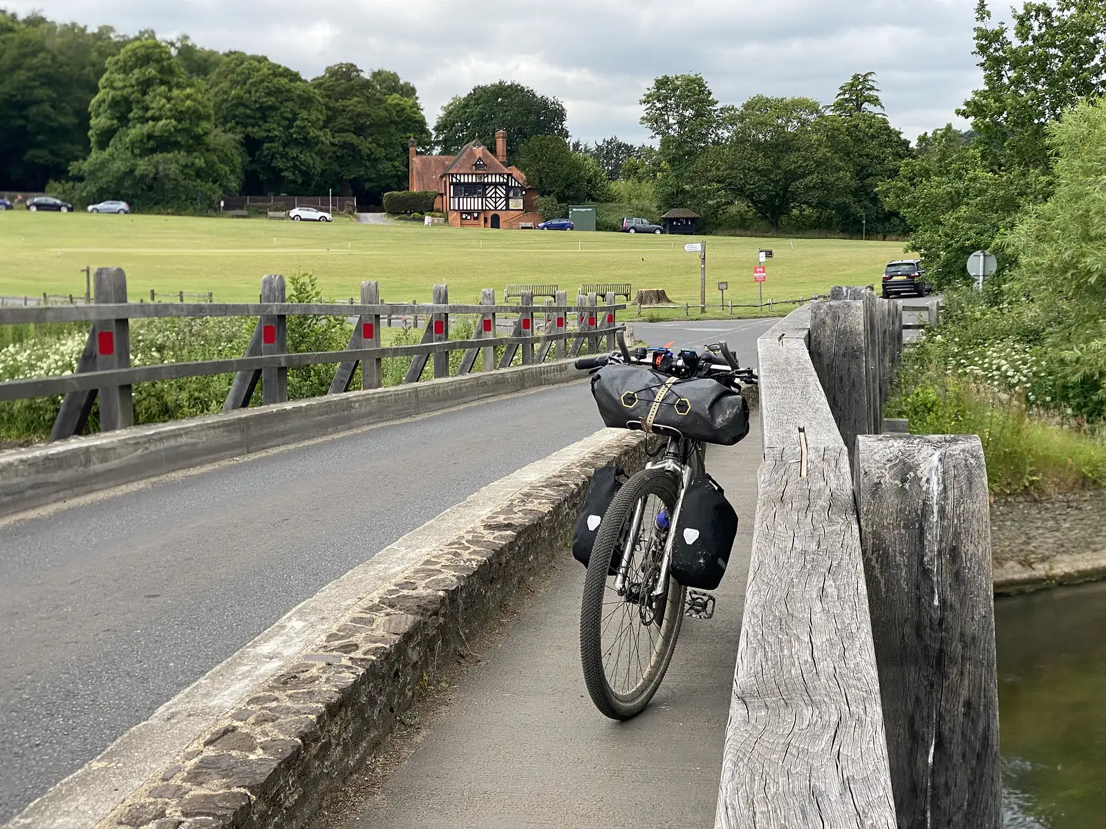 A loaded touring bike propped against the timber rail of a low bridge over a river, with a brick and half-timbered cricket pavilion across the green.