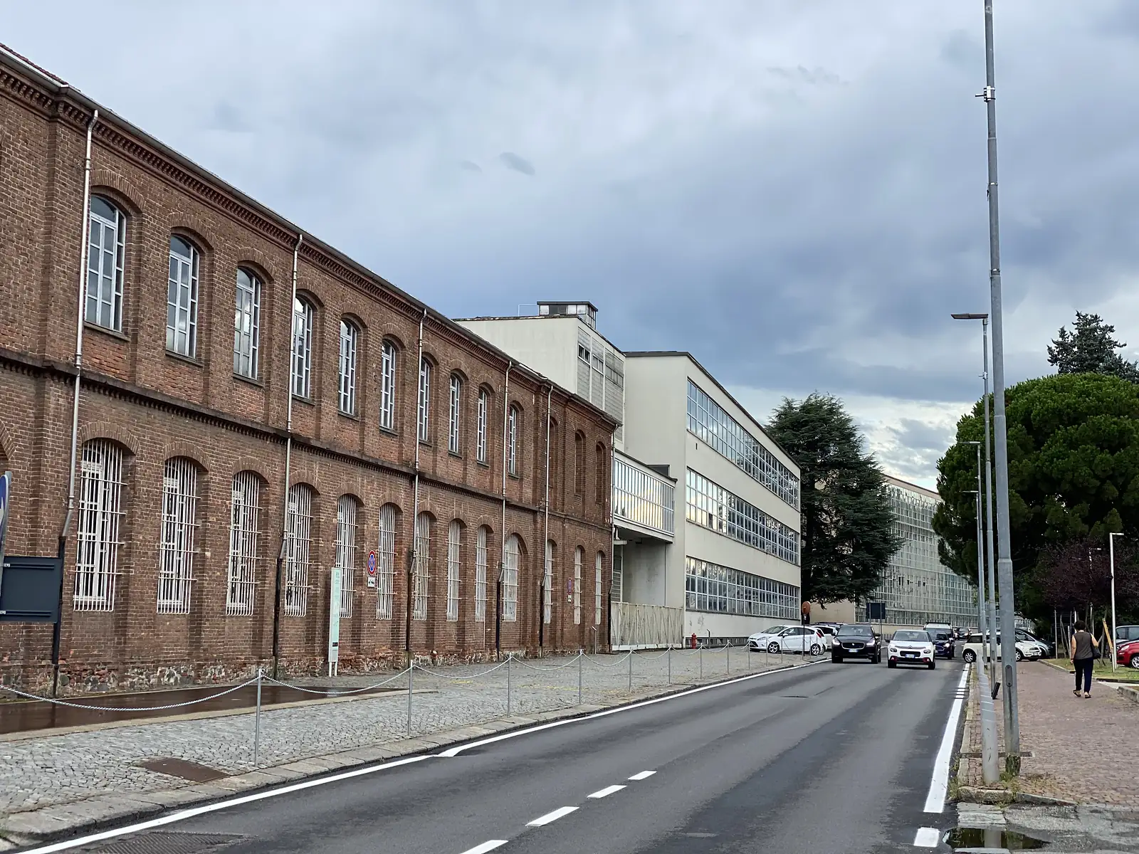 A long red-brick industrial building with arched, barred windows lining a street, with a plainer mid-century block visible further along.