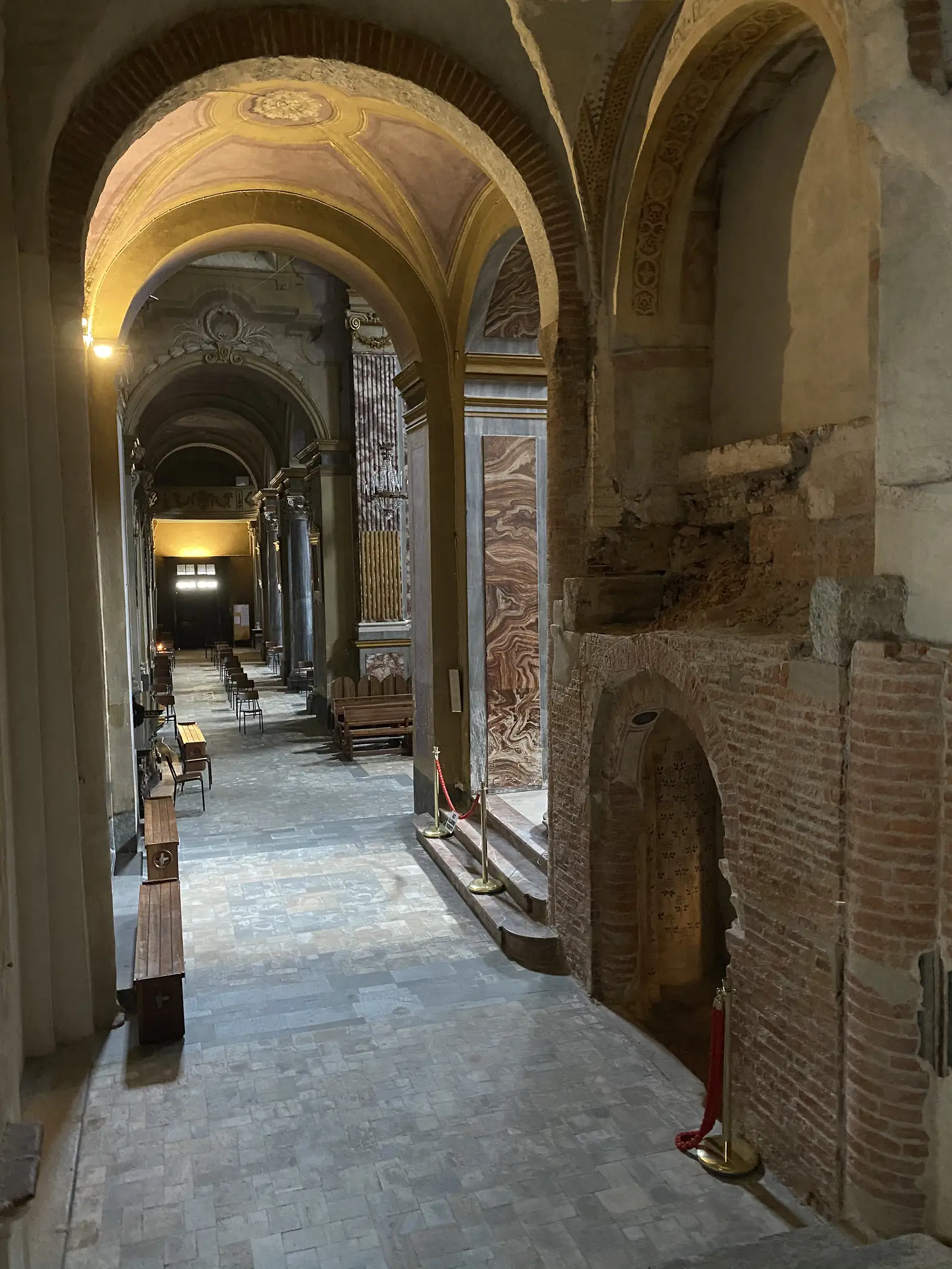A church side aisle with marbled pilasters and wooden benches, opening on the right onto a section of exposed medieval brickwork roped off from visitors.
