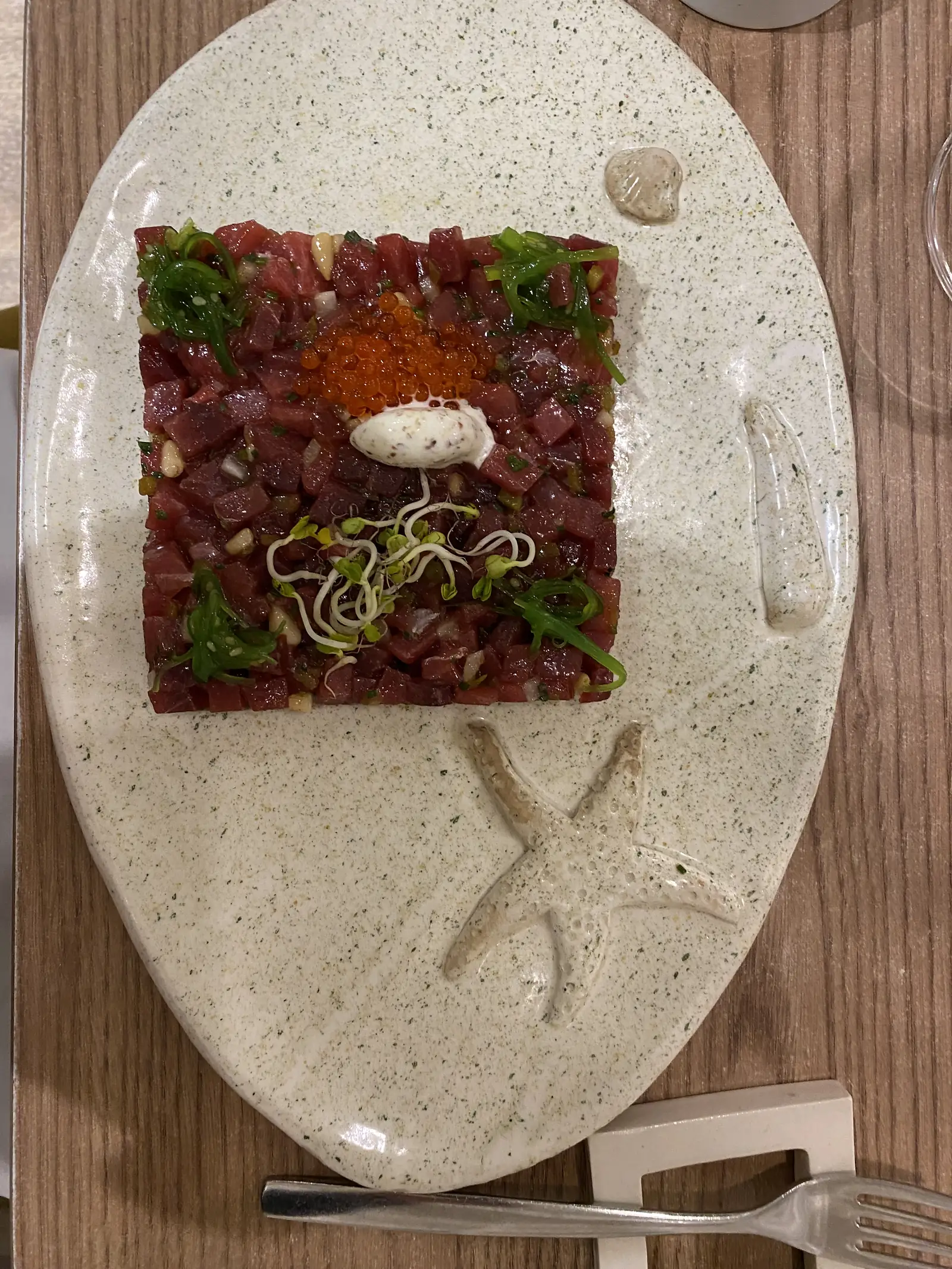 A square of diced red tuna tartare topped with orange roe, seaweed and sprouted seeds, on a speckled oval plate moulded with a starfish.