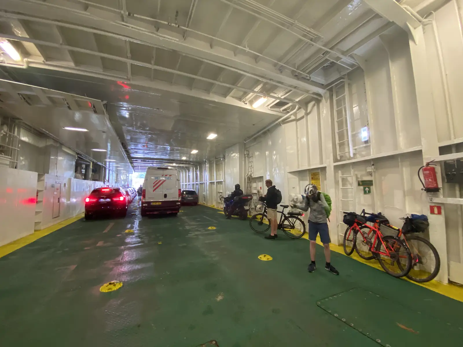 Cyclists and cars on a ferry's green-floored vehicle deck, loaded bikes propped along the wall.