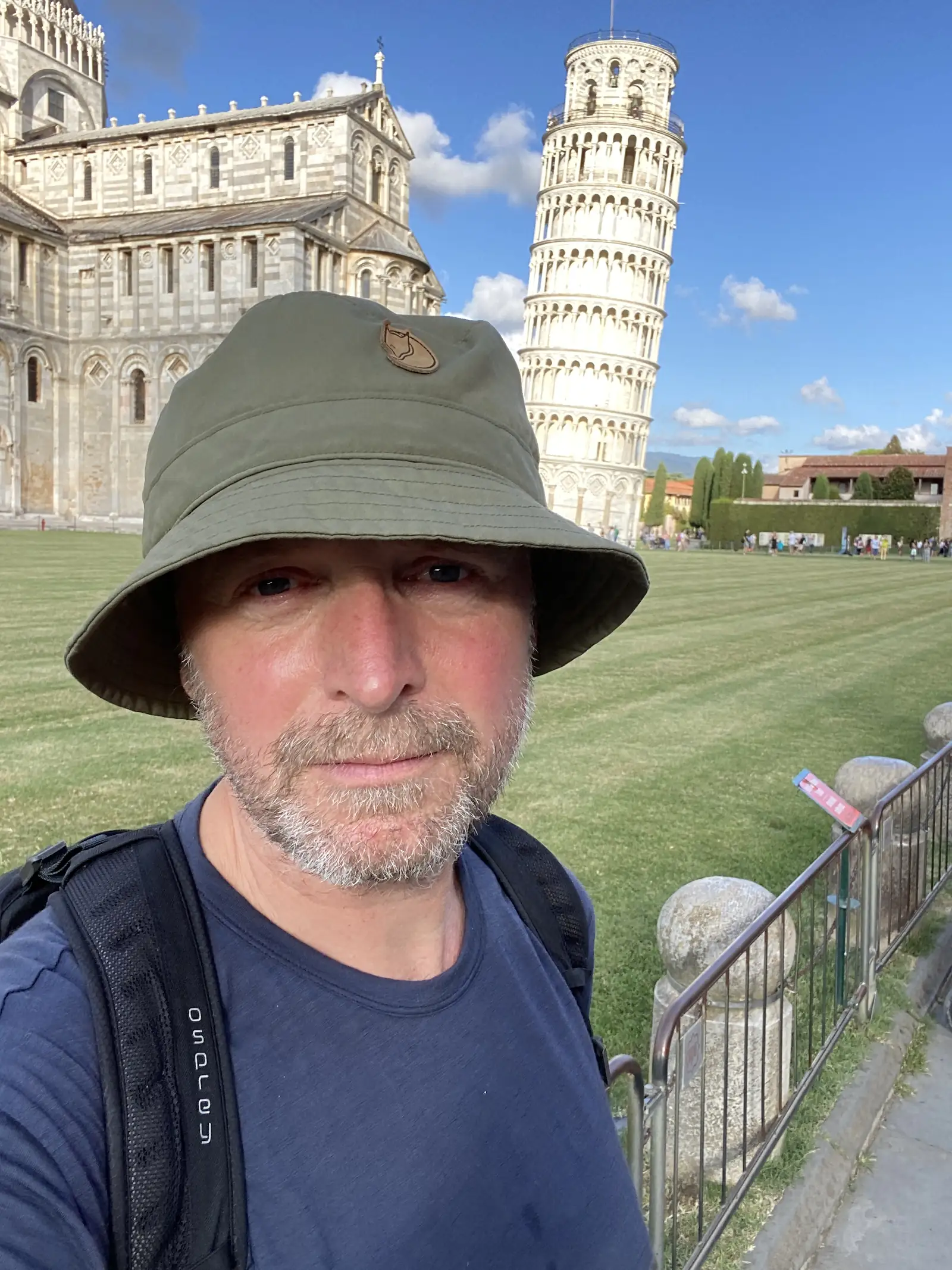 A self-portrait of a bearded man in a wide-brimmed hat and backpack, the Leaning Tower and cathedral visible on the grass behind him.