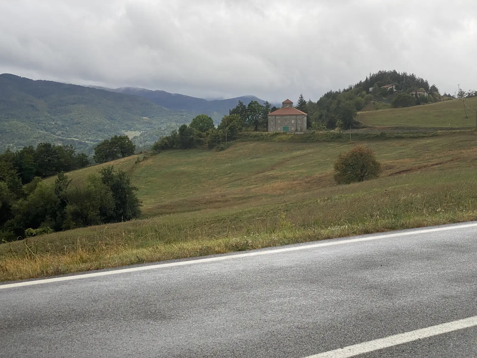 A square stone farmhouse with a pyramid-roofed tower and terracotta tiles sits alone on a grassy hillside, distant mountains fading into low cloud behind it.