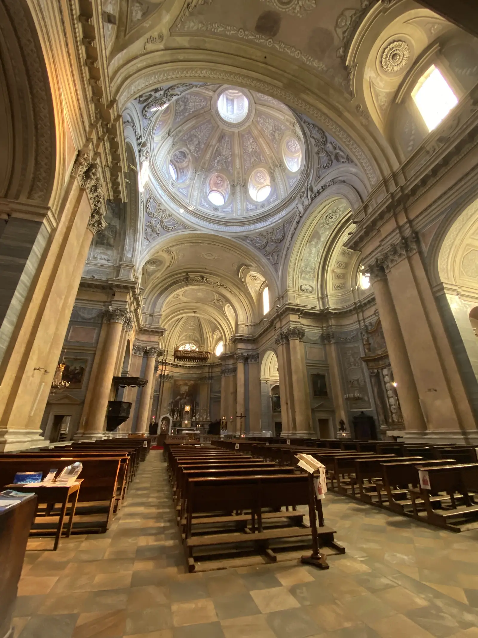 The ornately painted domed ceiling and nave of a baroque church, with wooden pews lining the aisle below.