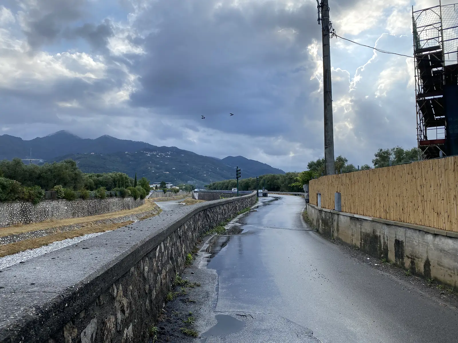 An empty tarmac lane runs beside a stone retaining wall above a dry riverbed, with distant hills, a bamboo screen fence and construction scaffolding under a cloudy sky with two birds overhead.