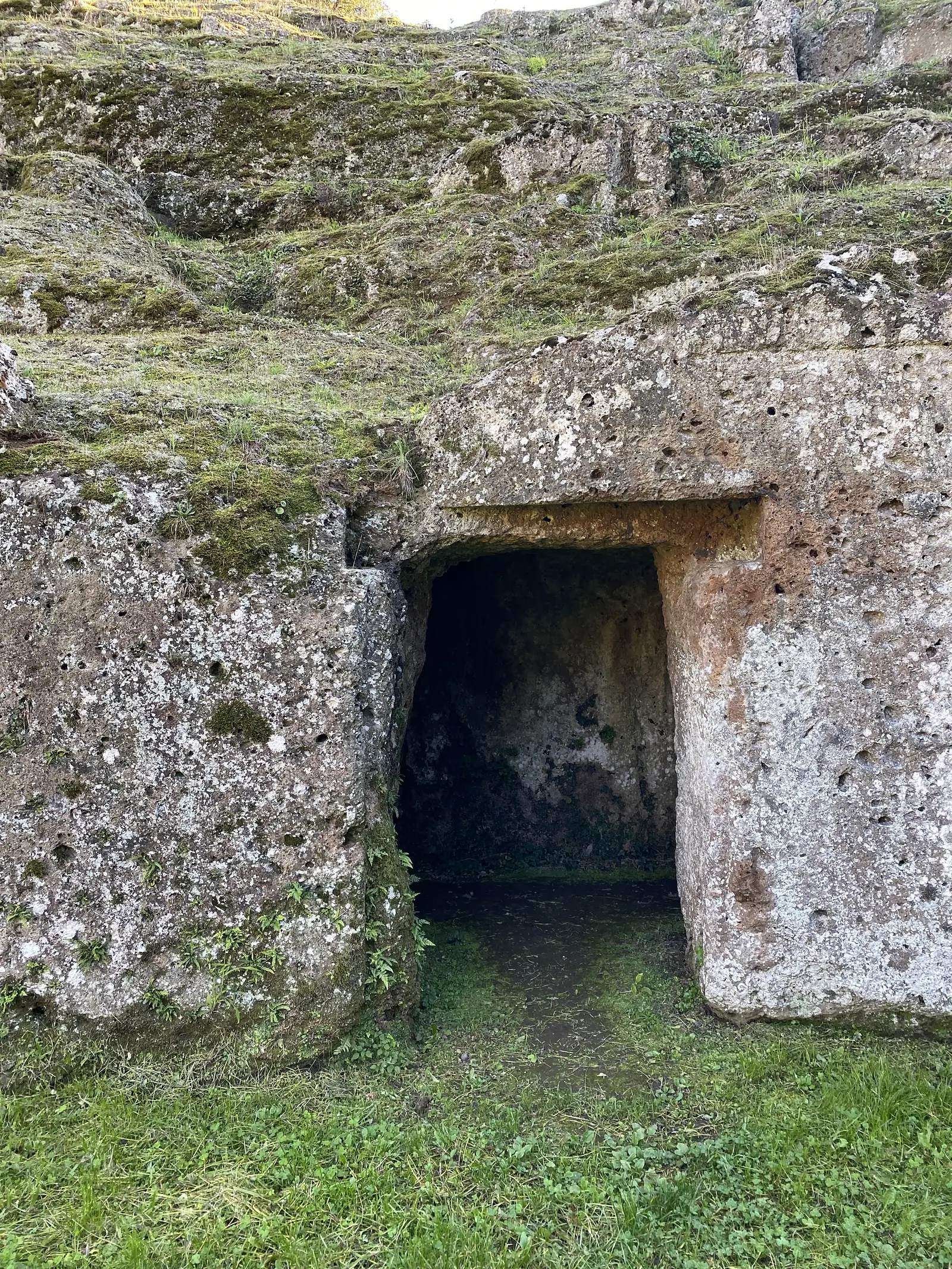 A small rectangular niche cut into an ancient tufa rock-face, likely a burial recess, with grass growing at its edge.