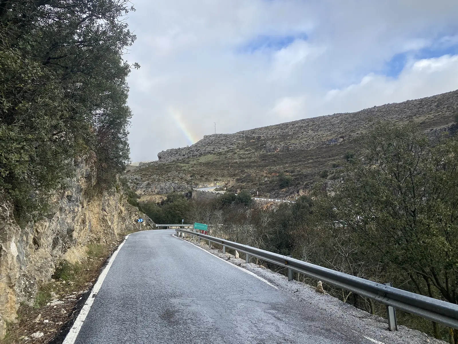 A wet road curving beneath a rock cutting, with a faint rainbow standing over the bare hillside beyond the crash barrier.