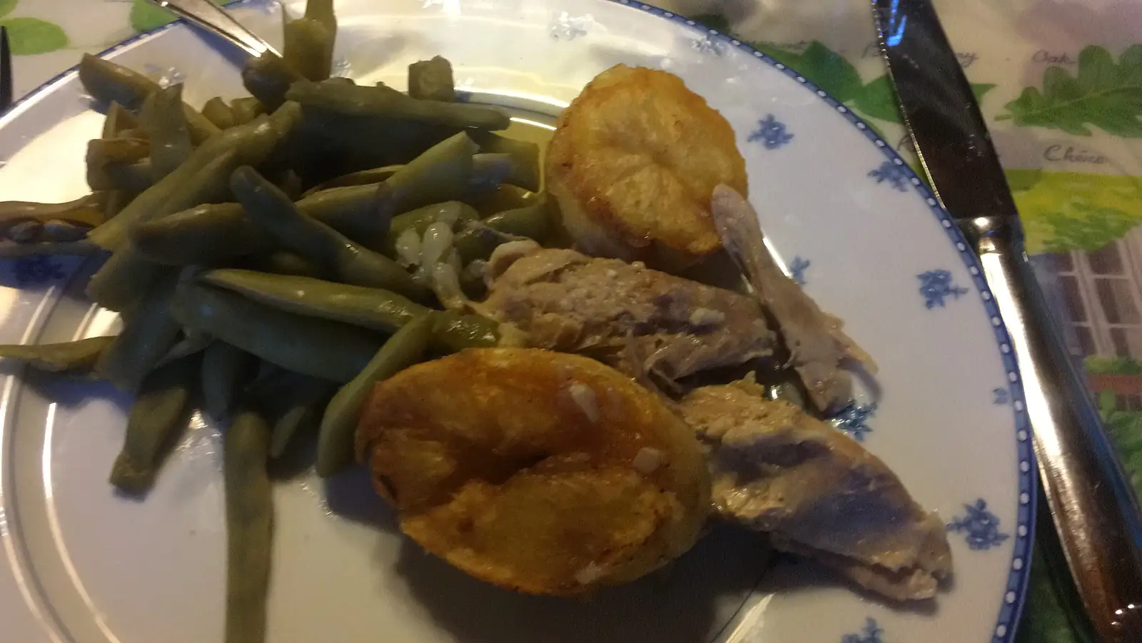 A dinner plate of green beans, shallots and roasted potato halves alongside pieces of roast game, set on a floral tablecloth beside a knife and fork.