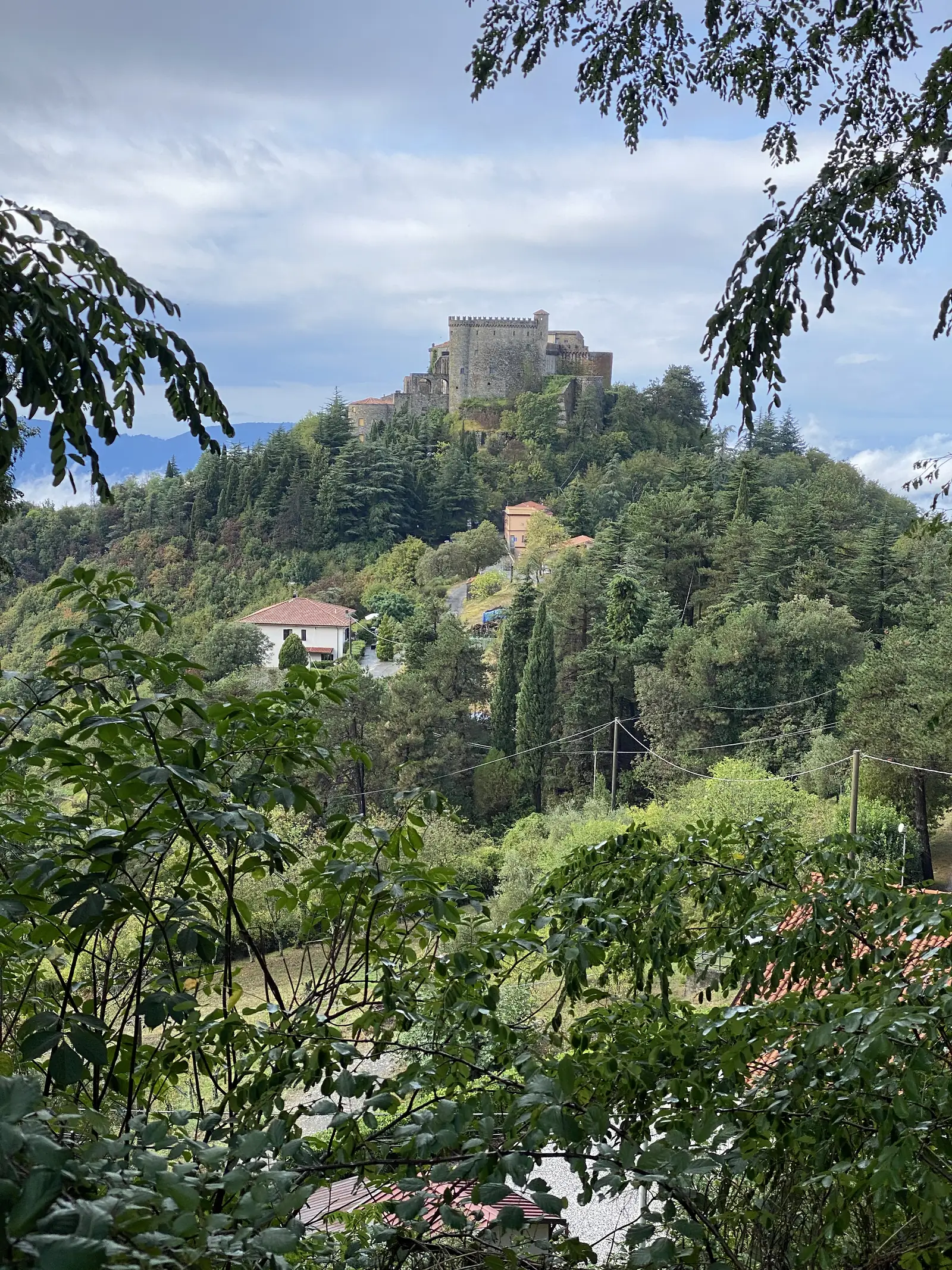 A stone tower with castellated battlements rising above a cluster of village rooftops and tall cypress trees, seen across the valley under a partly cloudy sky.