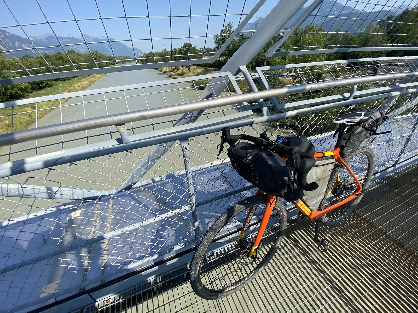 A laden orange gravel bike rests against the mesh railing of a pedestrian bridge over a straight river channel, mountains visible in the distance.
