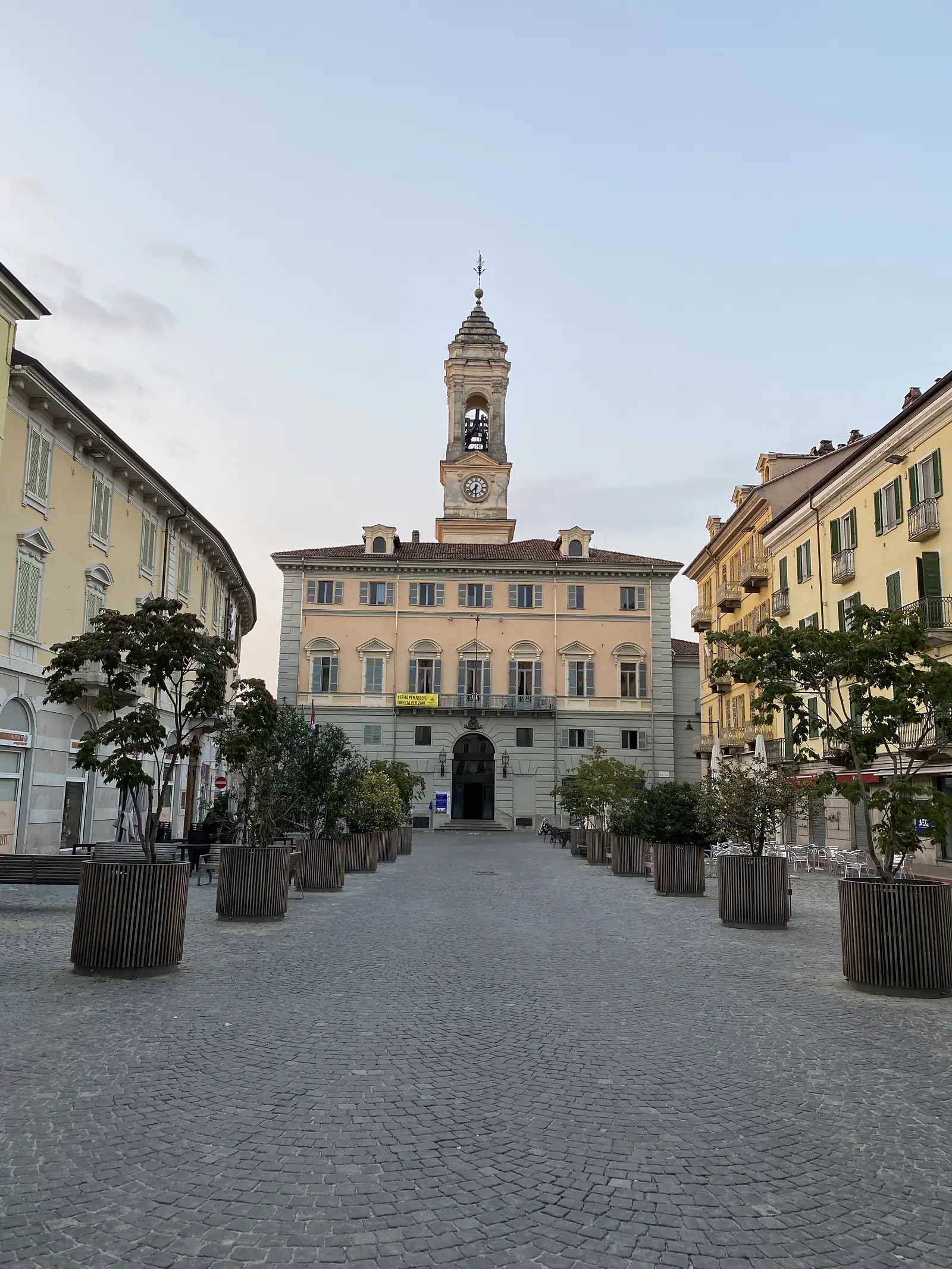 A tiered clock tower rises above a pink and cream piazza building, with café tables and umbrellas set out on the cobbles below.