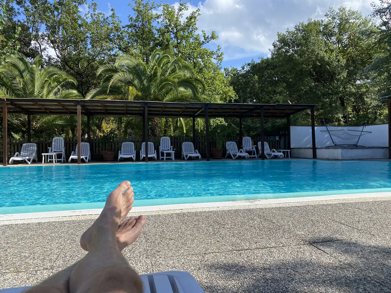 A pair of bare feet raised in the foreground of a swimming pool terrace lined with white sun loungers under a pergola, palm trees rising behind.