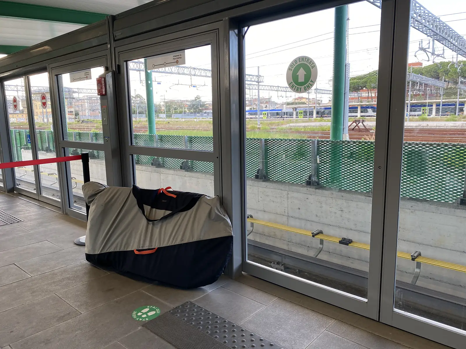 A covered walkway at the automated people-mover station signed 'Stazione FS Pisa Centro' and 'Solo Entrata Entry Only', with a padded bike bag propped against the glass wall and trains visible on the tracks beyond.
