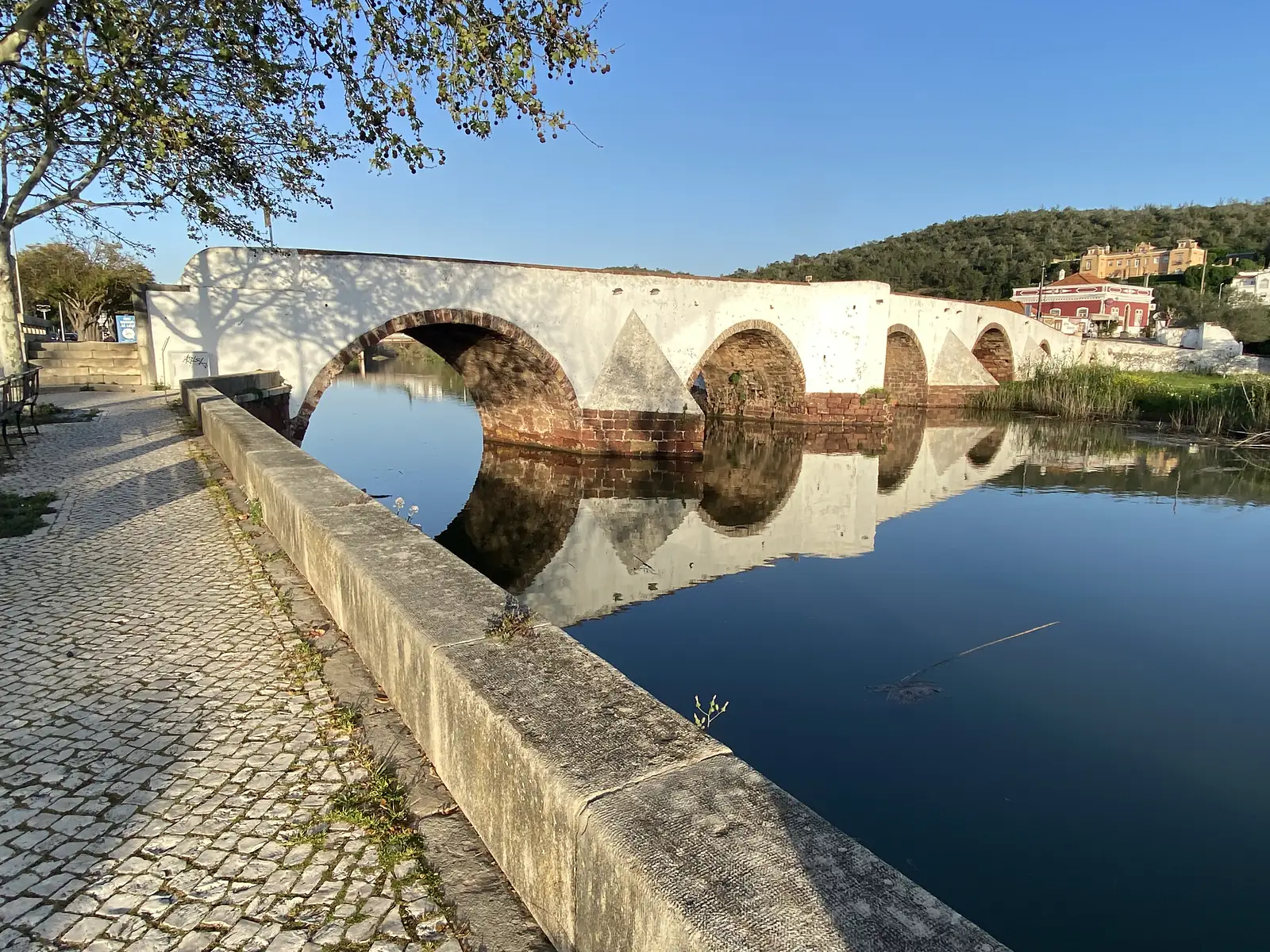 Whitewashed multi-arched stone bridge with red brick arch rings, mirrored perfectly in the still river below, a paved riverside walkway running along the near bank.