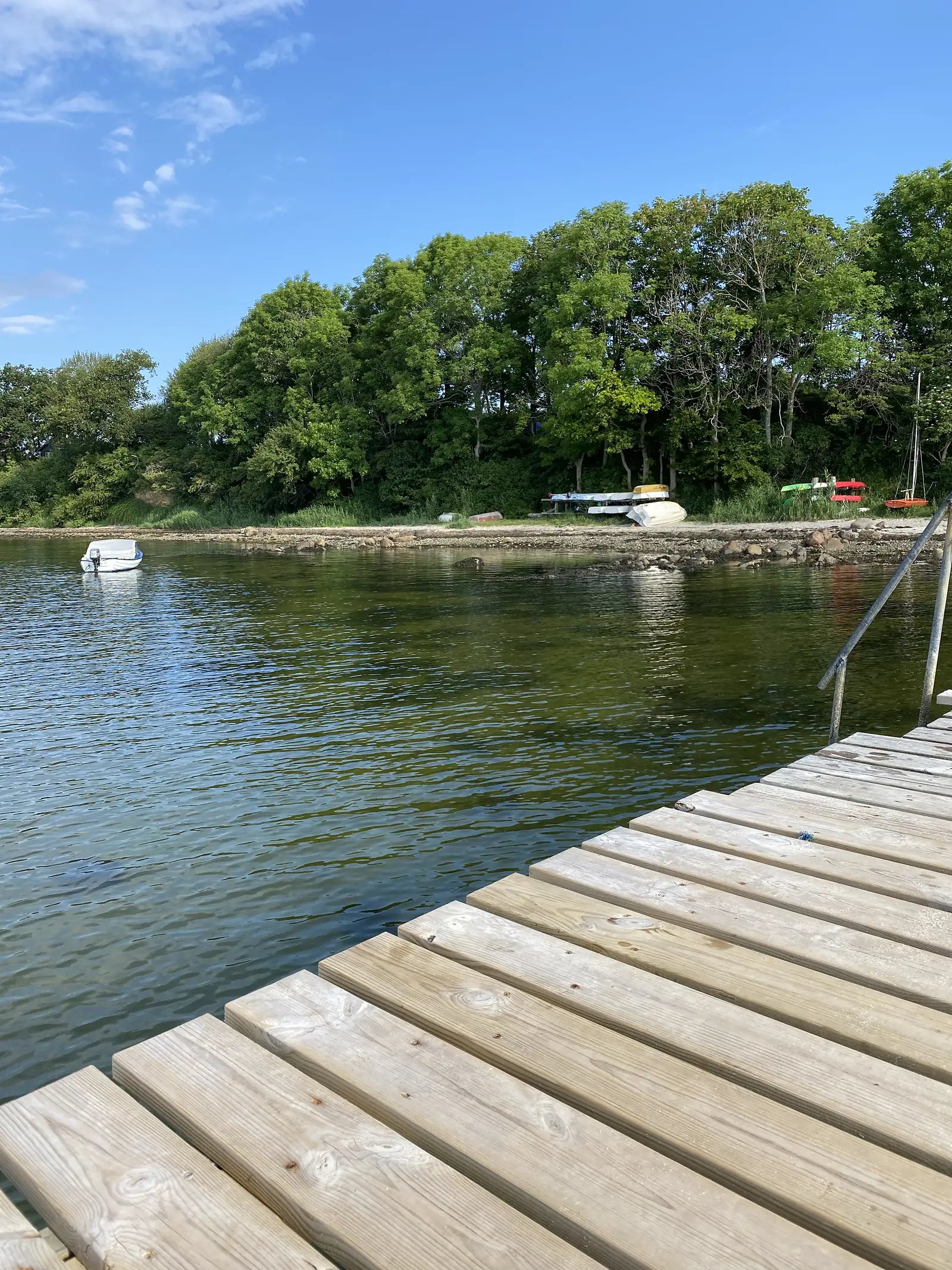 A wooden jetty running out into a calm blue inlet, with a small white boat moored and dinghies and kayaks drawn up on the wooded far shore.