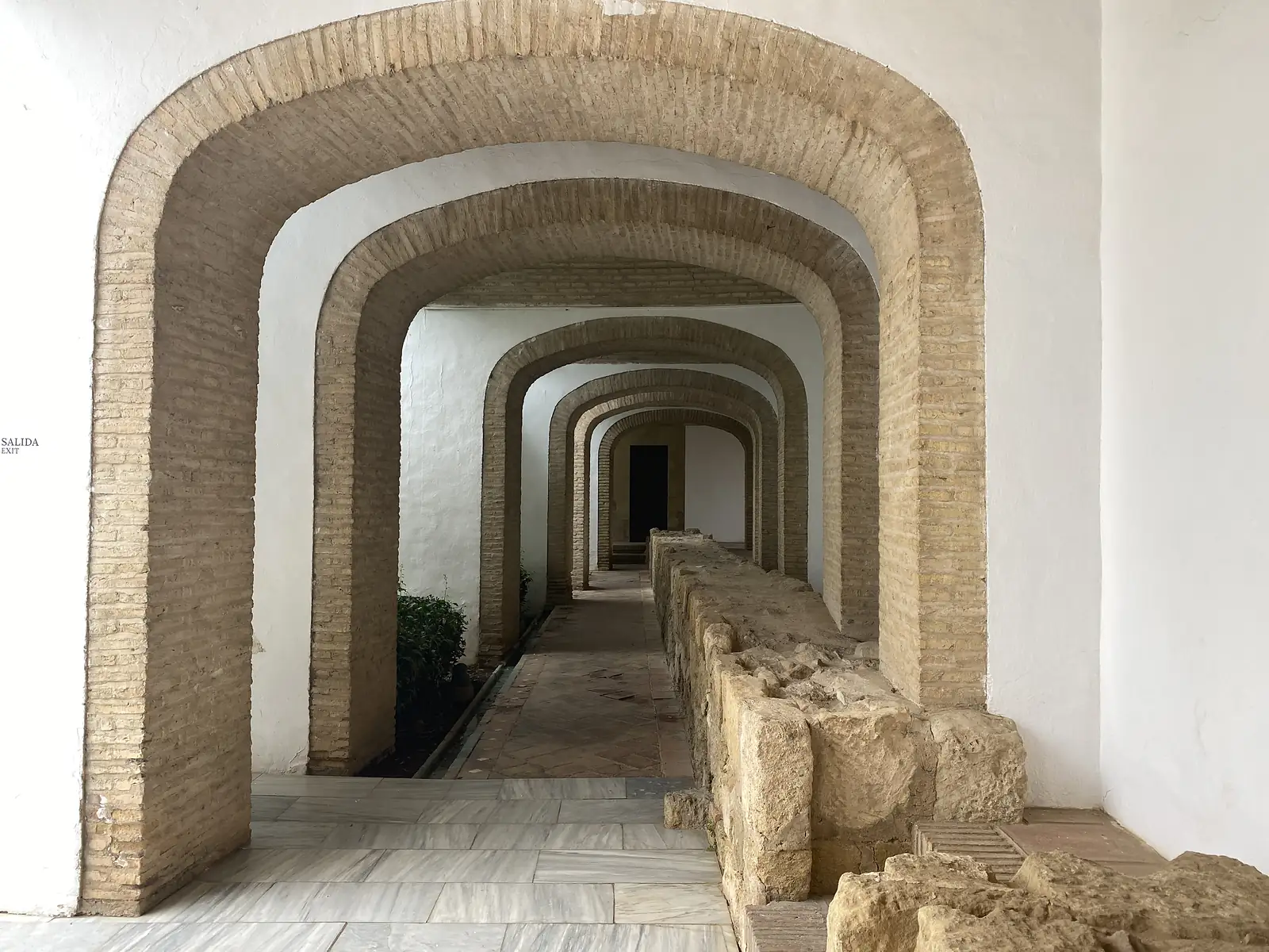 A run of receding brick horseshoe arches over a paved passage, excavated stone foundations alongside and a sign reading "SALIDA / EXIT".
