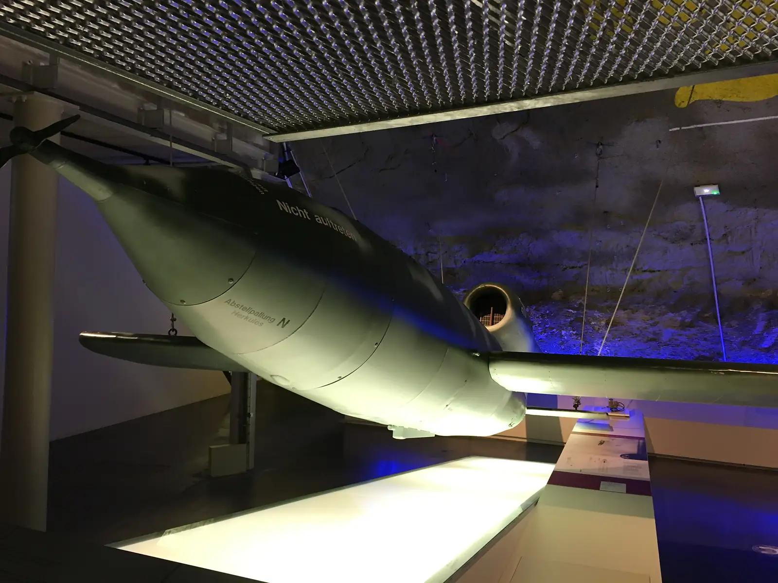A green and white flying-bomb replica with a propeller nose and the stencilled warning 'Nicht auftreten' hangs beneath a wire-mesh ceiling, lit in blue.