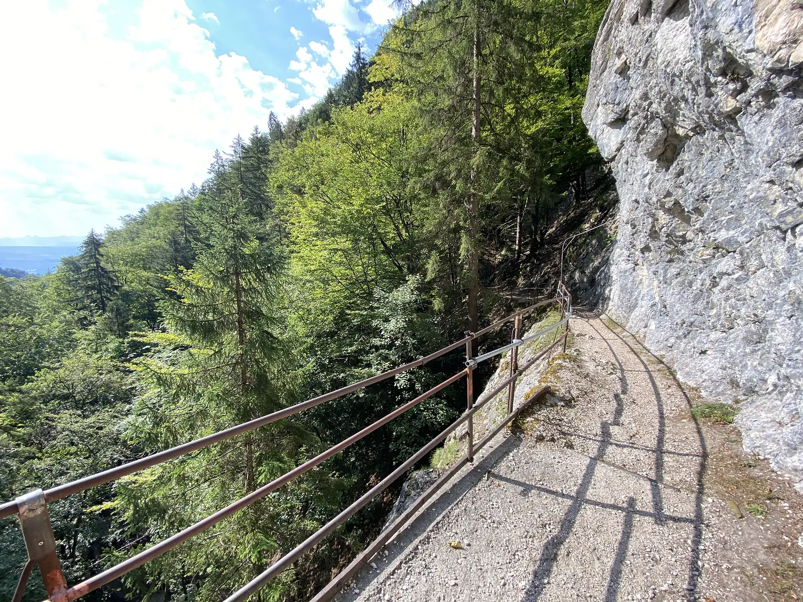 A gravel path skirting a sheer limestone cliff on a wooded hillside, with a rusted iron handrail and hazy hills visible through the trees below.