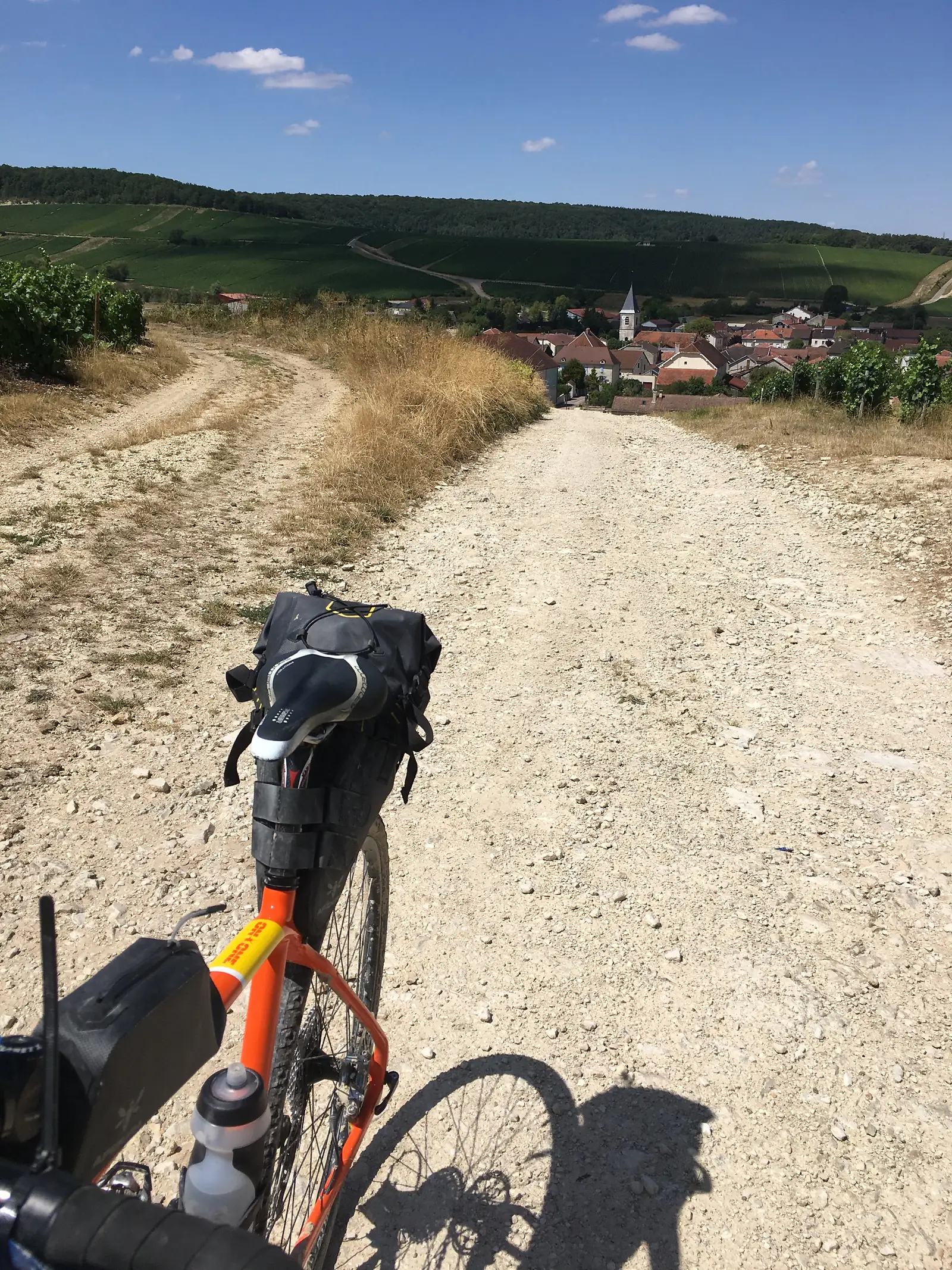 A bikepacking saddle bag and sunglasses sit atop a bicycle seat, with a chalky white track leading down towards a village in the distance.