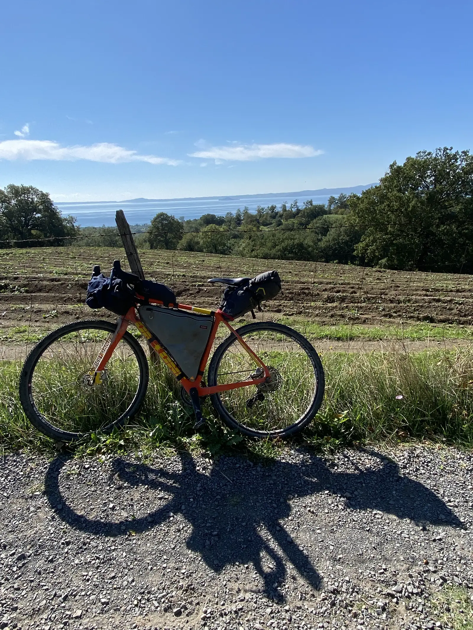 A loaded orange bikepacking bike stands on a gravel track beside a fenced field, its rider's shadow cast across the stones.
