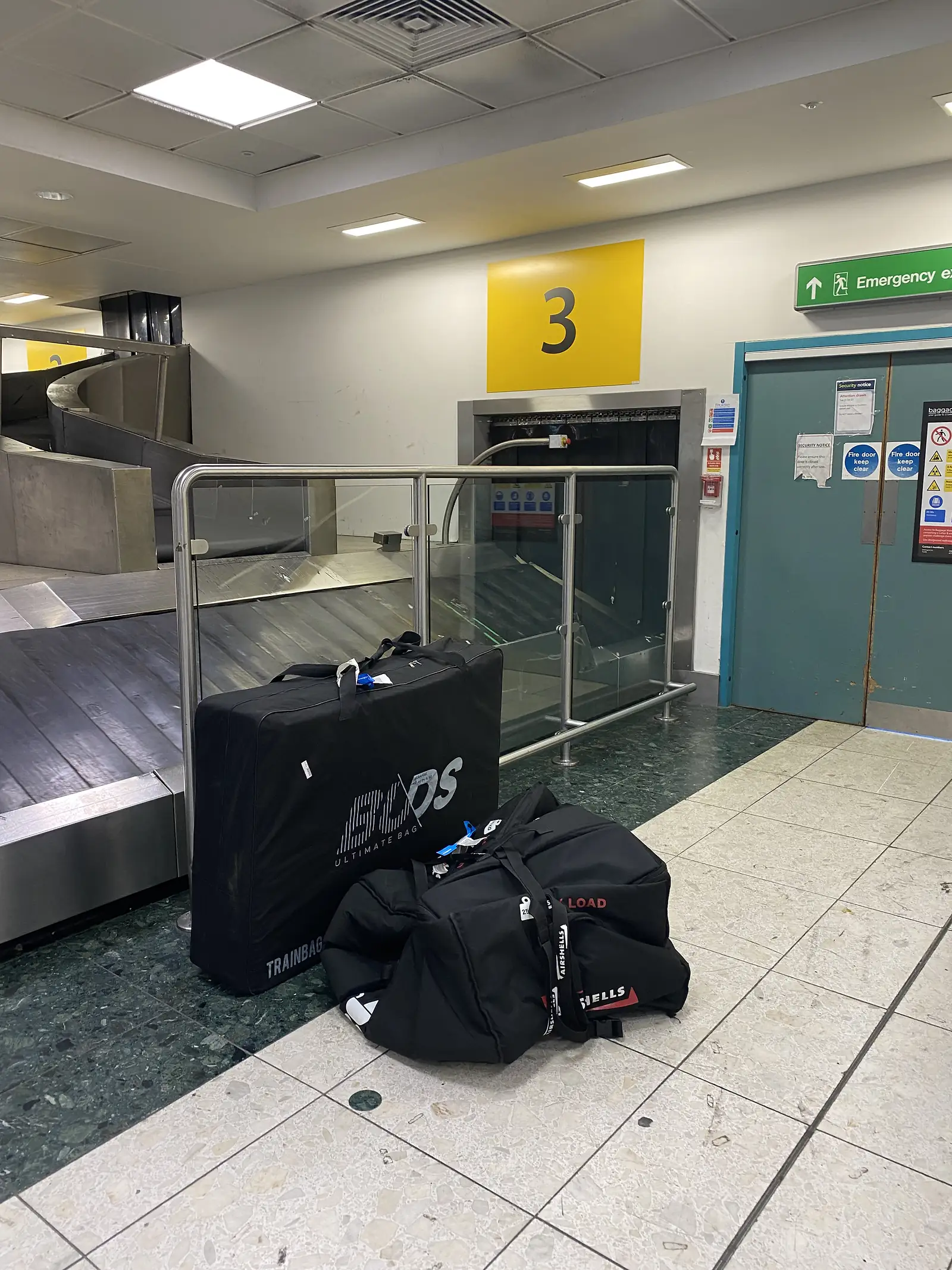 A padded bike travel bag and a black holdall on the floor beside baggage carousel 3 in an airport arrivals hall.
