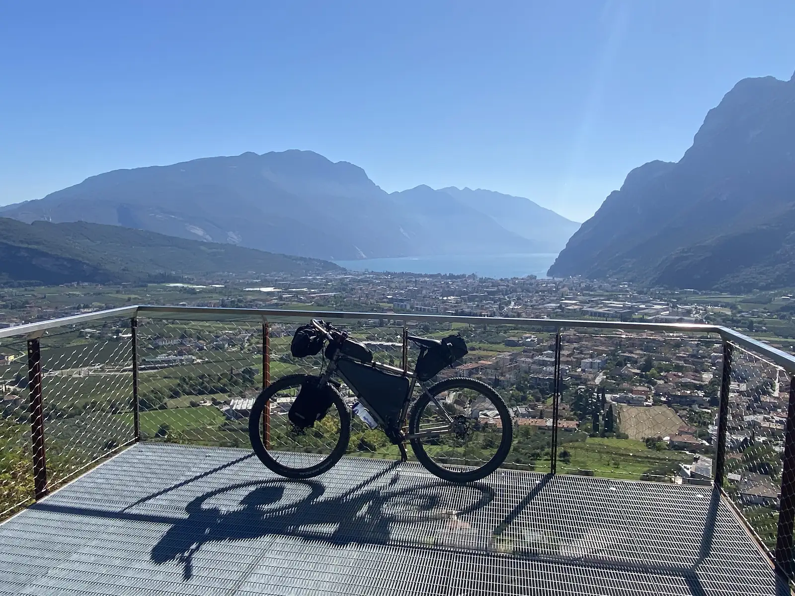 A loaded bikepacking bike stands on a steel viewing platform high above a town at the head of a lake, with mountain walls on either side.
