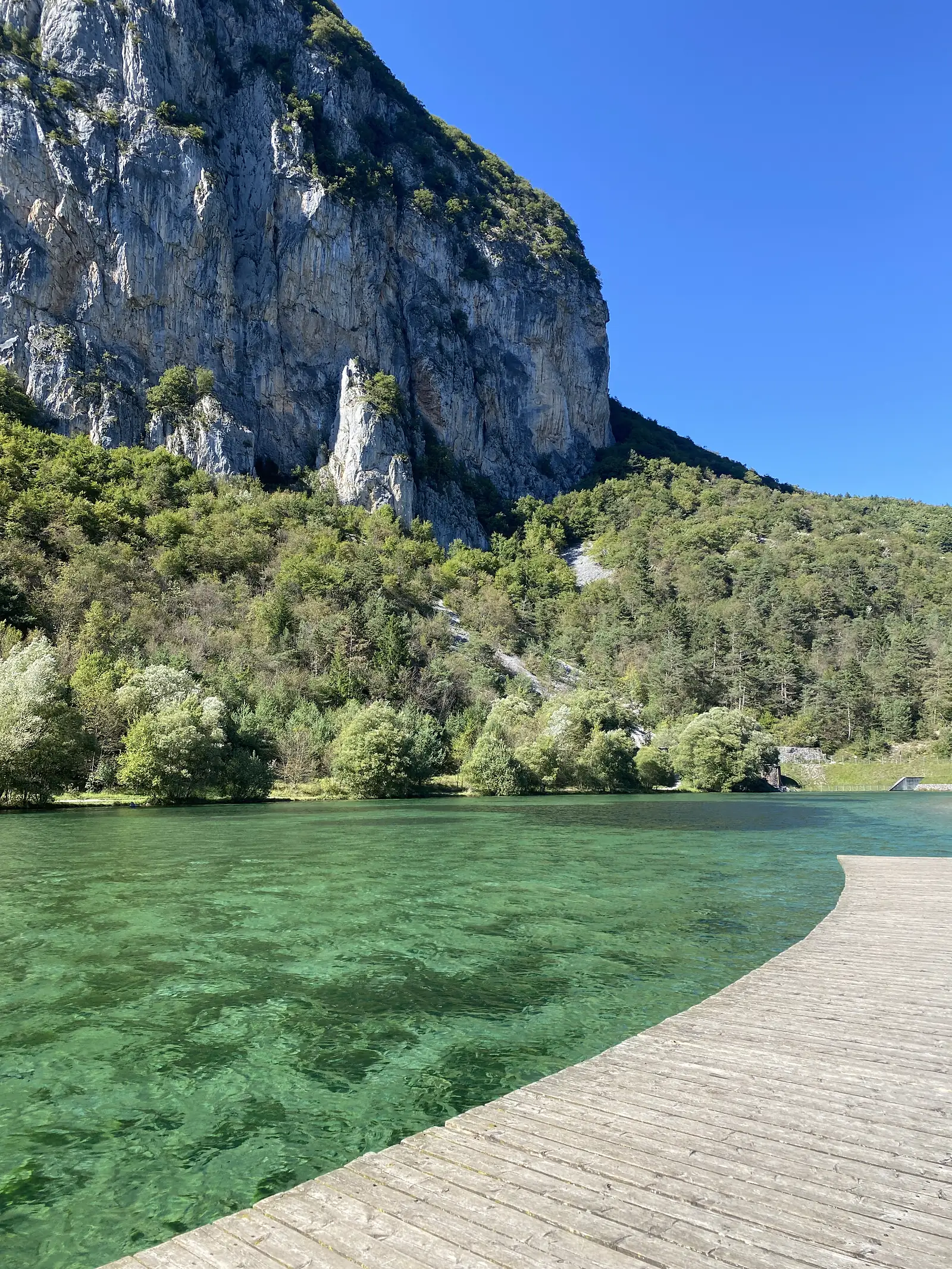 A wooden boardwalk curves along the edge of a clear green lake beneath a towering limestone cliff.