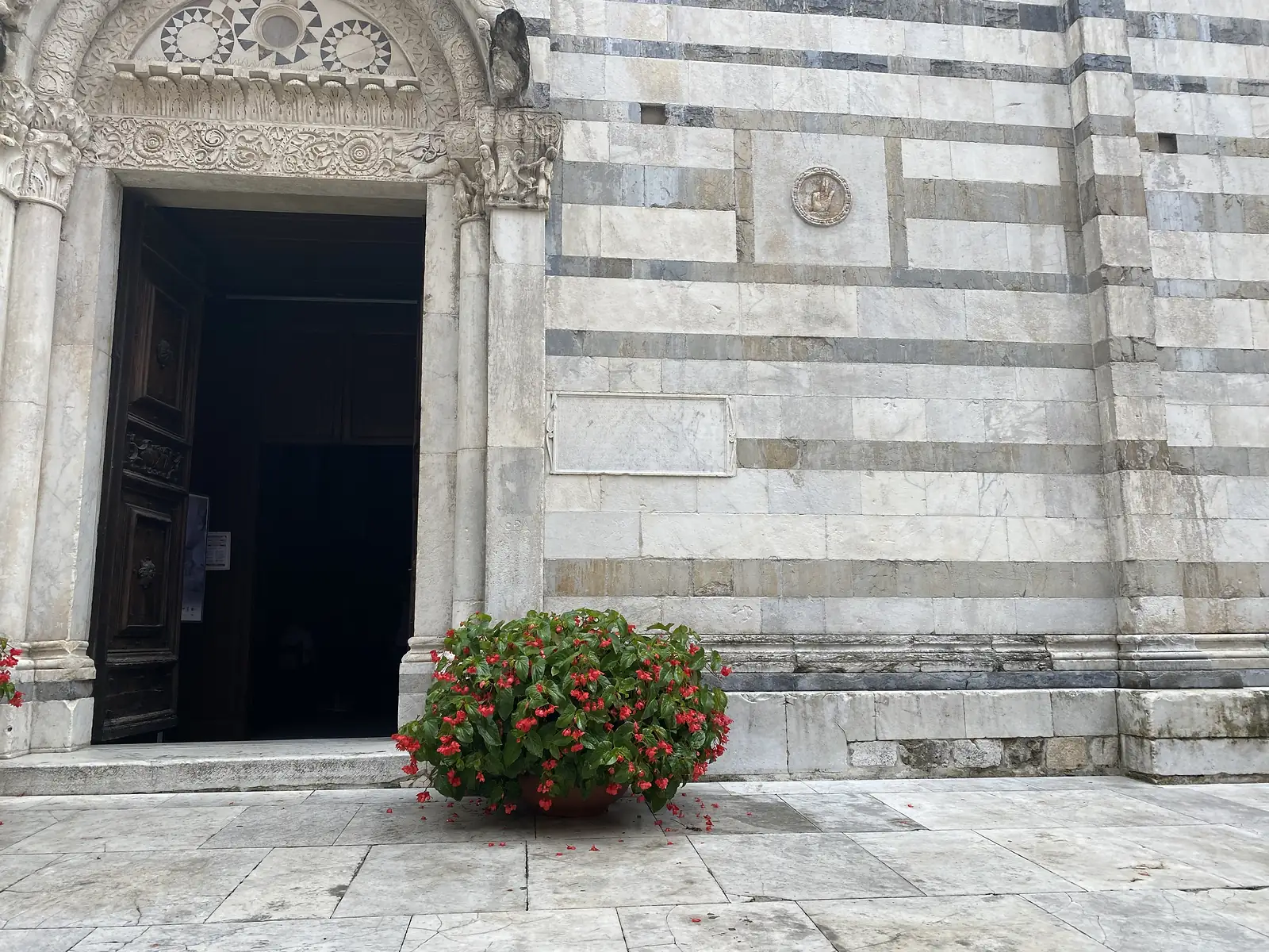 The carved marble doorway of a striped marble church, with a stone plaque set into the wall and a pot of bright red begonias beside the open dark wood door.