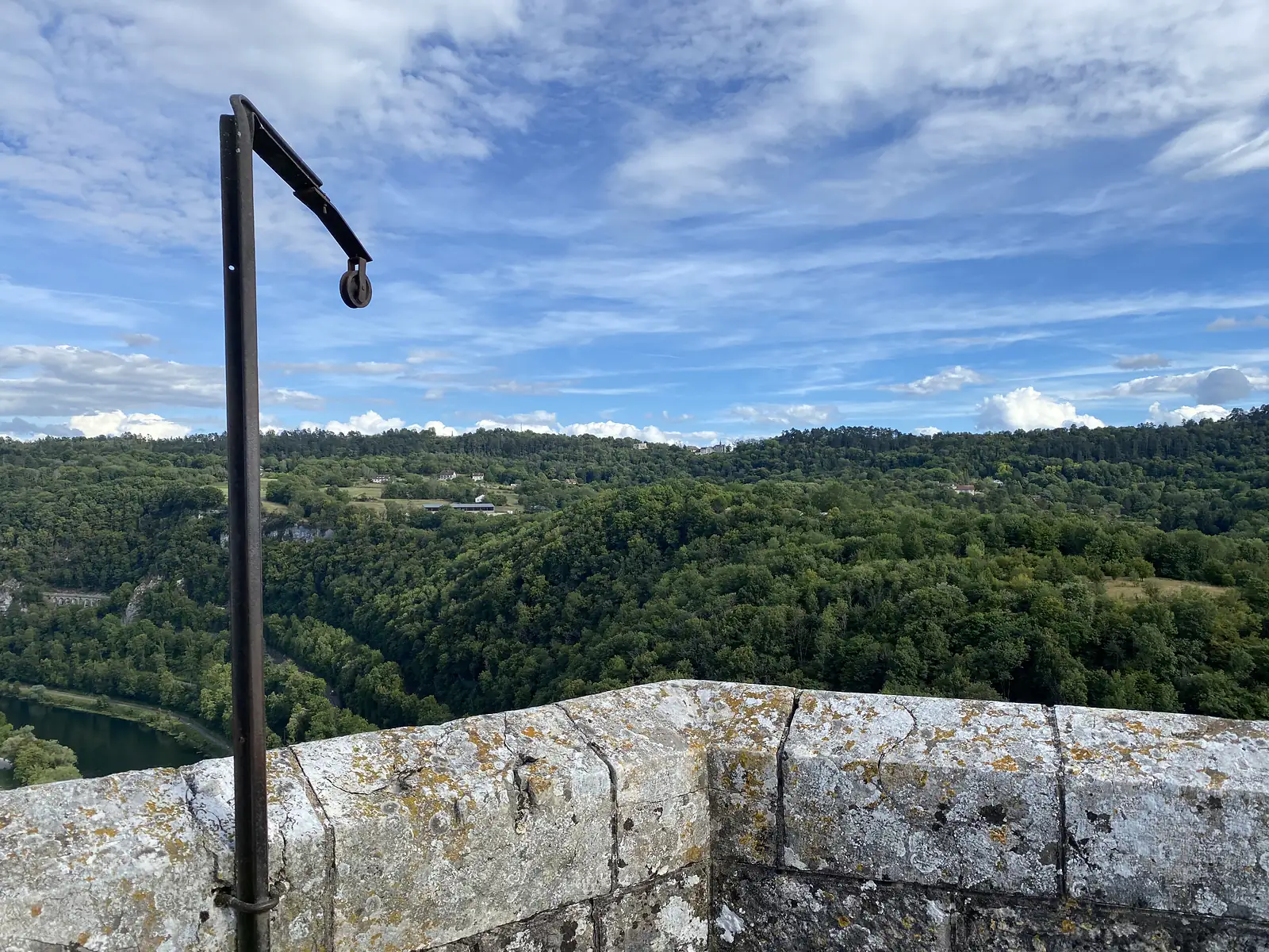 A weathered iron hoist with a pulley wheel stands on a lichen-covered rampart, overlooking a wooded river gorge.