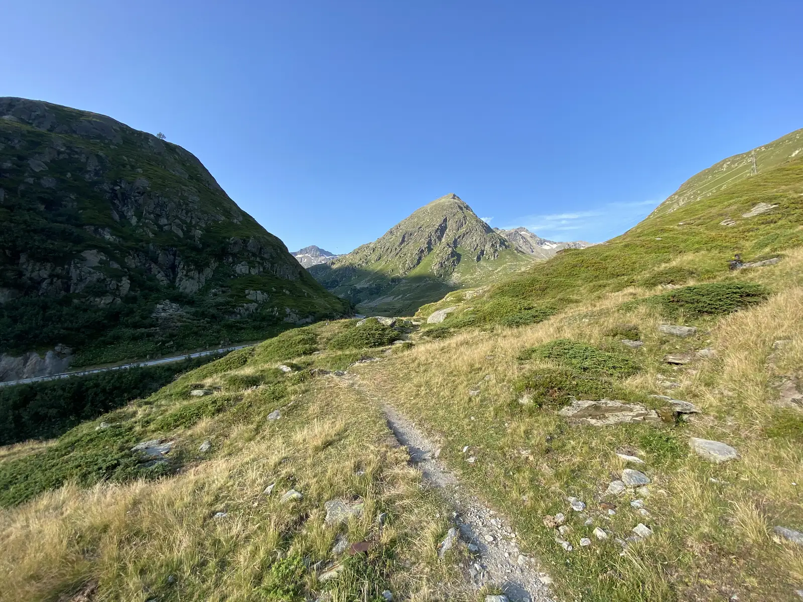 A stony trail winds down a grassy slope towards a hairpin road bend, with layered mountain ridges beyond.