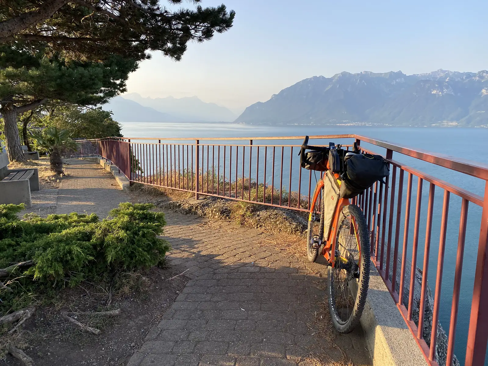 A loaded orange bikepacking bike leaning against a red railing at a lakeside viewpoint, overlooking Lake Geneva and the Alps in early morning light.