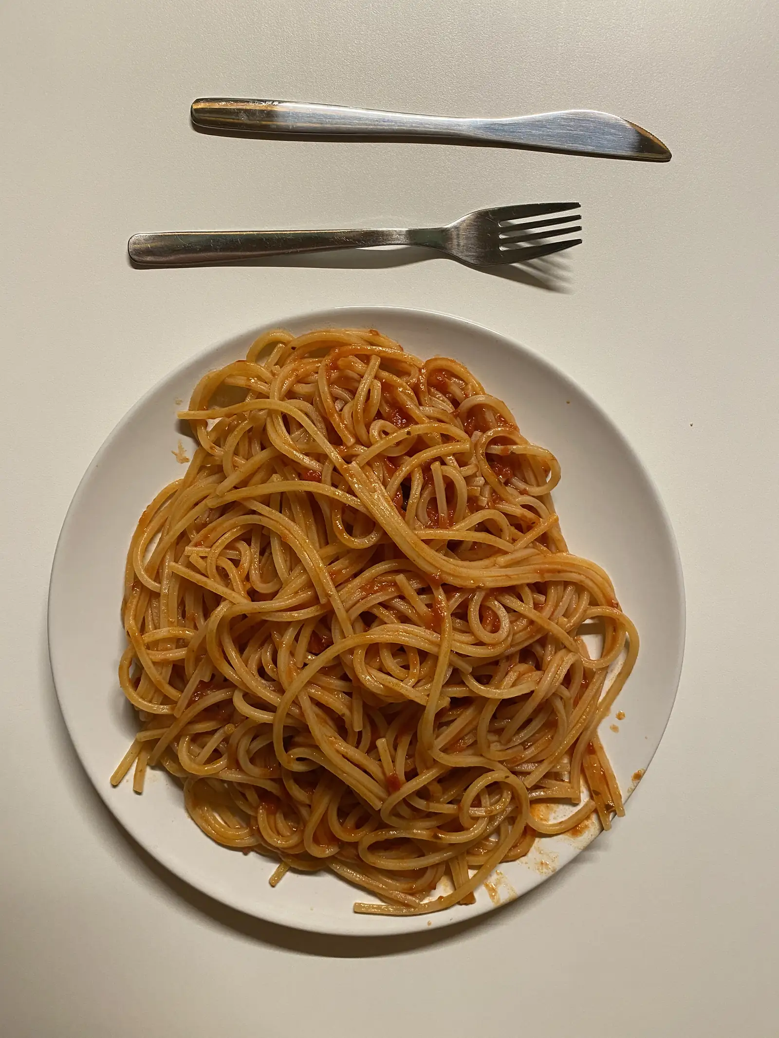 A plate of spaghetti in tomato sauce sits on a table beside a knife and fork.