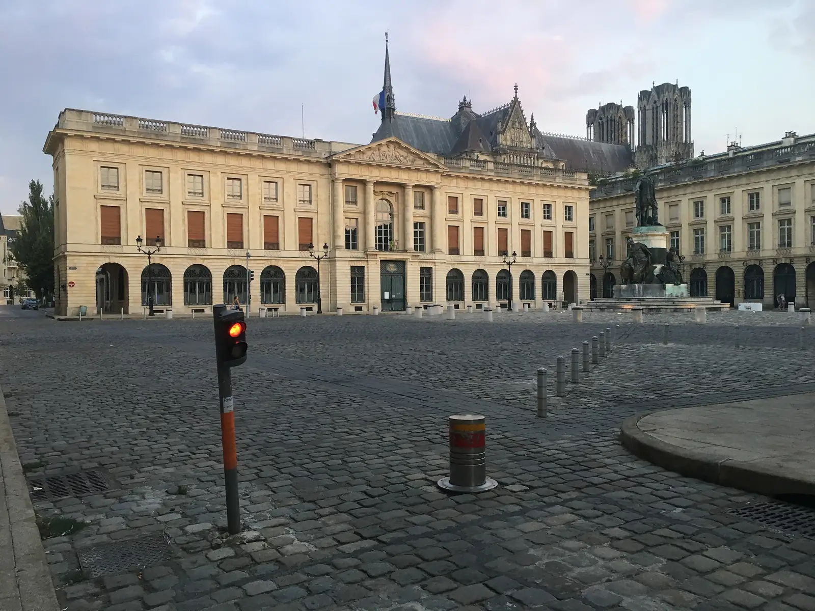 A cobbled square with a lit red traffic signal and a bollard stands before a grand colonnaded building, with Reims cathedral's towers rising behind at dusk.