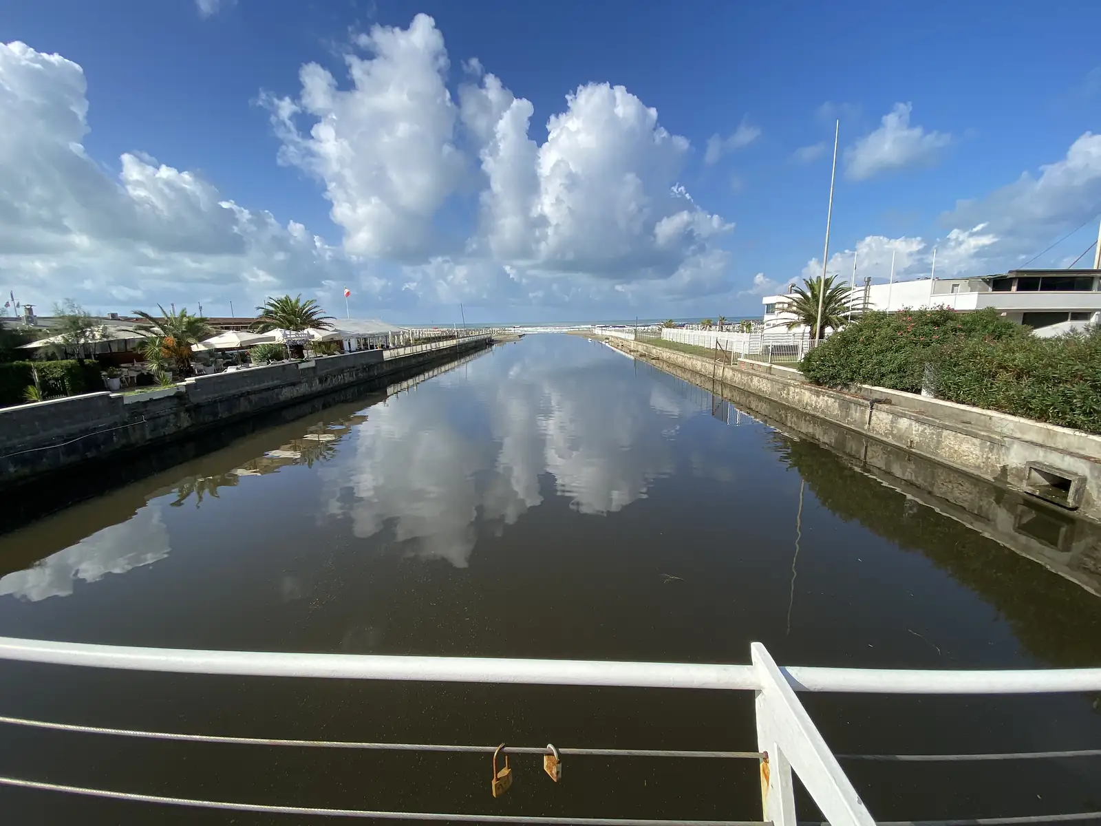 Two padlocks are fixed to a bridge railing overlooking a still canal that reflects clouds and palm trees, with seafront buildings and the sea visible at the far end.