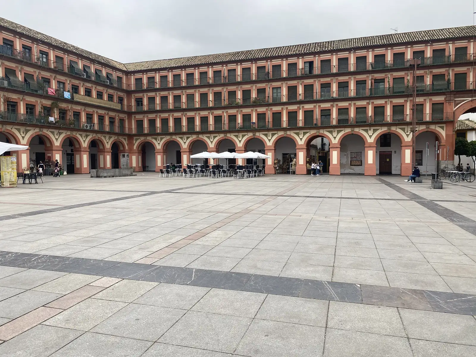 A large arcaded square ringed by four storeys of terracotta-and-cream balconied facades, with cafe tables under white parasols beneath the arches.