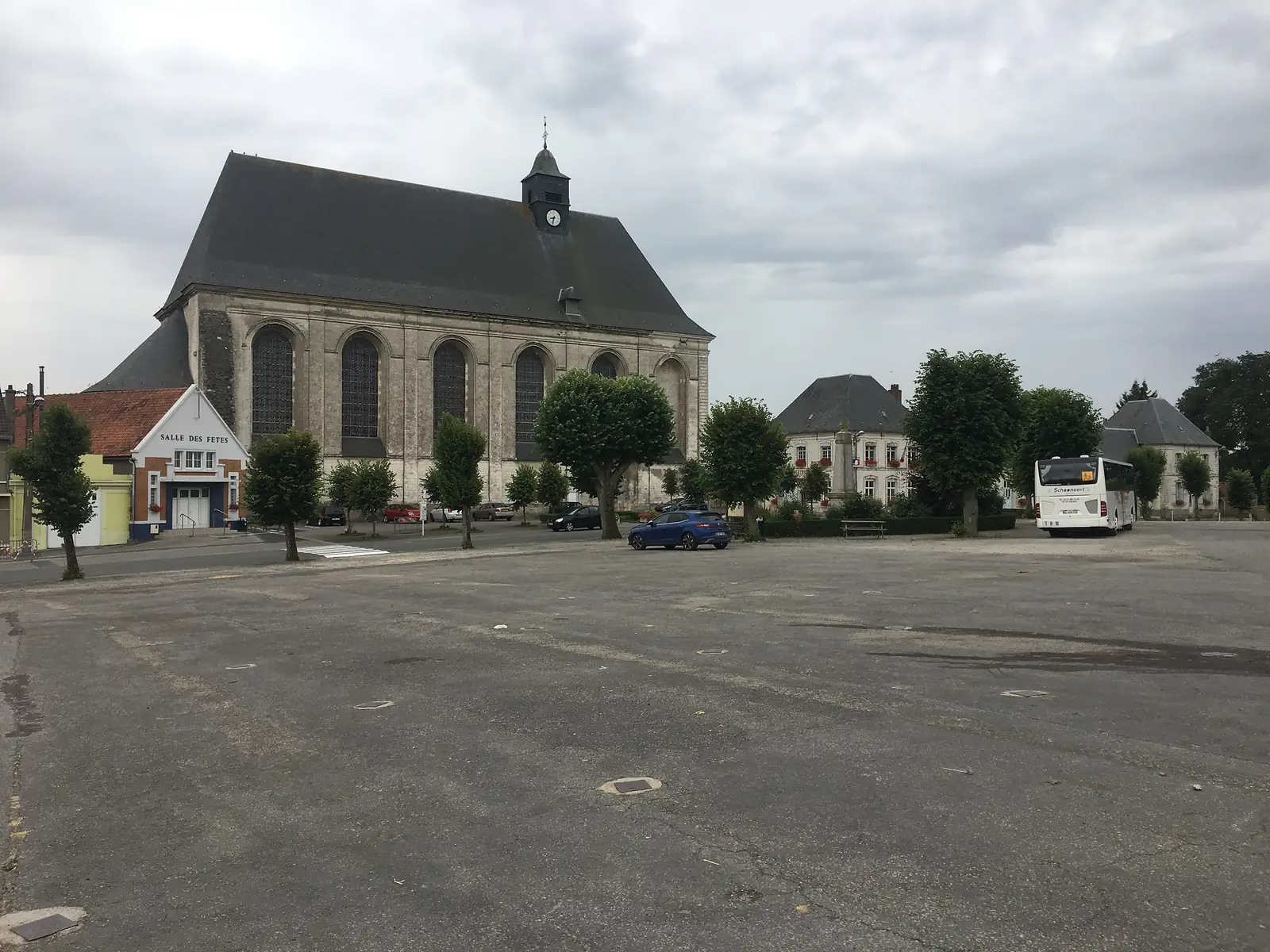 A stone church with a slate roof and clock tower stands beside a near-empty town square, with a coach, parked cars and the 'Salle des Fêtes' hall nearby.