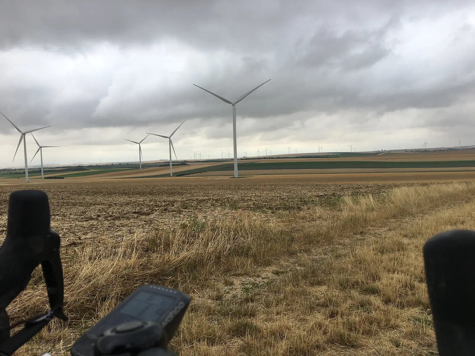 Wind turbines rise from rolling farmland, photographed past the dark shapes of a bicycle saddle and GPS unit in the foreground.