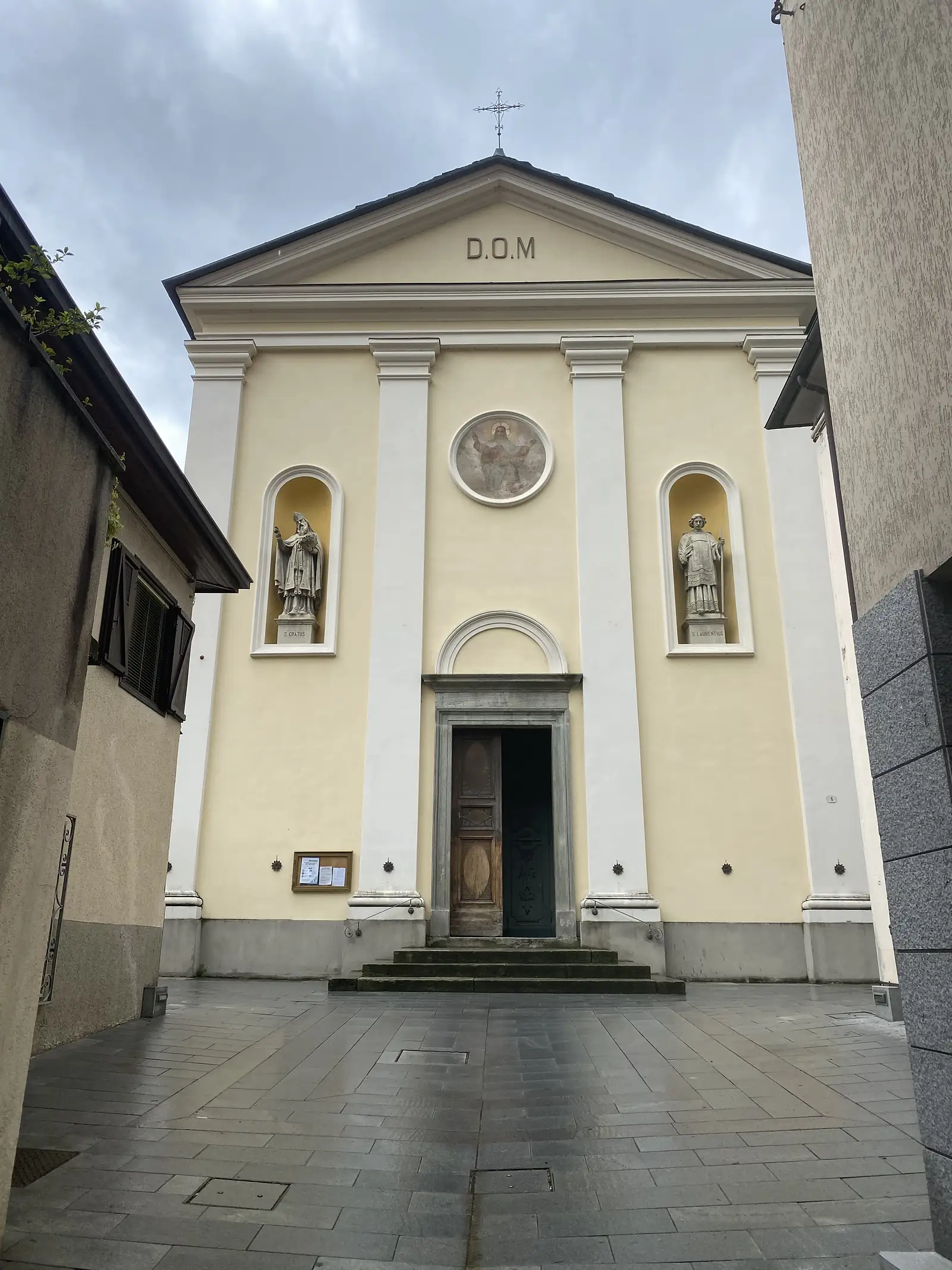 A pale yellow church facade with 'D.O.M' carved above the pediment, statues of saints in niches either side of a circular fresco, and worn stone steps leading up to its open wooden door.