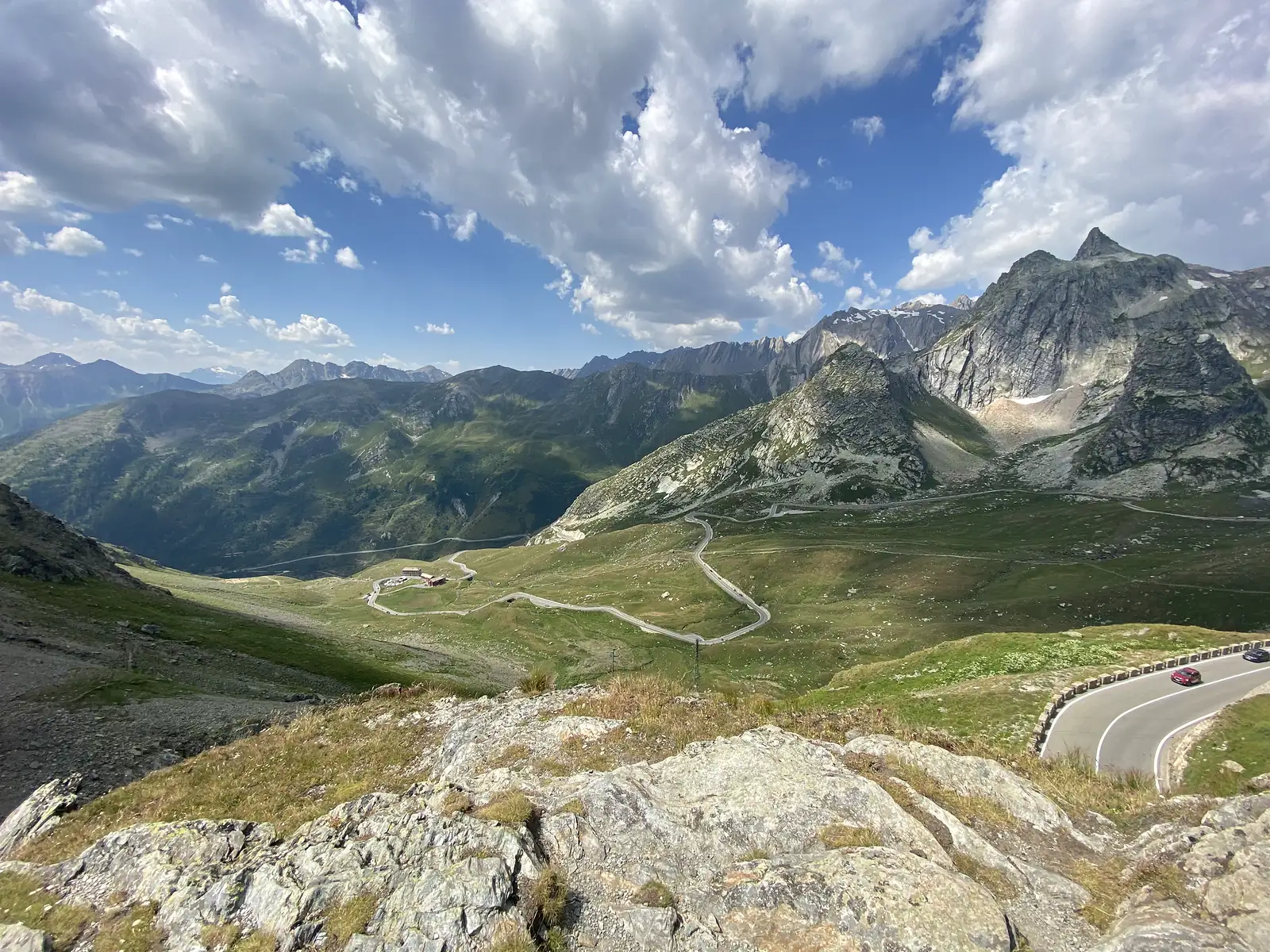 A hairpin mountain road winding through a green alpine valley below jagged rocky peaks, with a car rounding the nearest bend.
