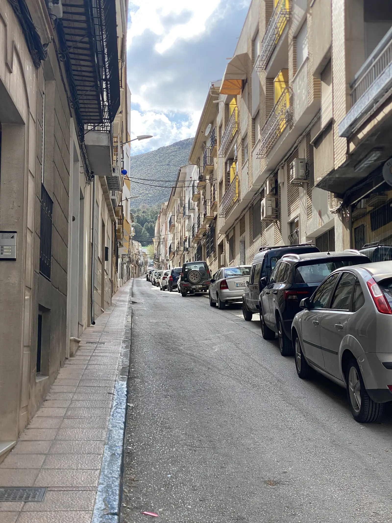 A narrow street of tall apartment blocks with cars parked along one side, a wooded mountain filling the end of the view.