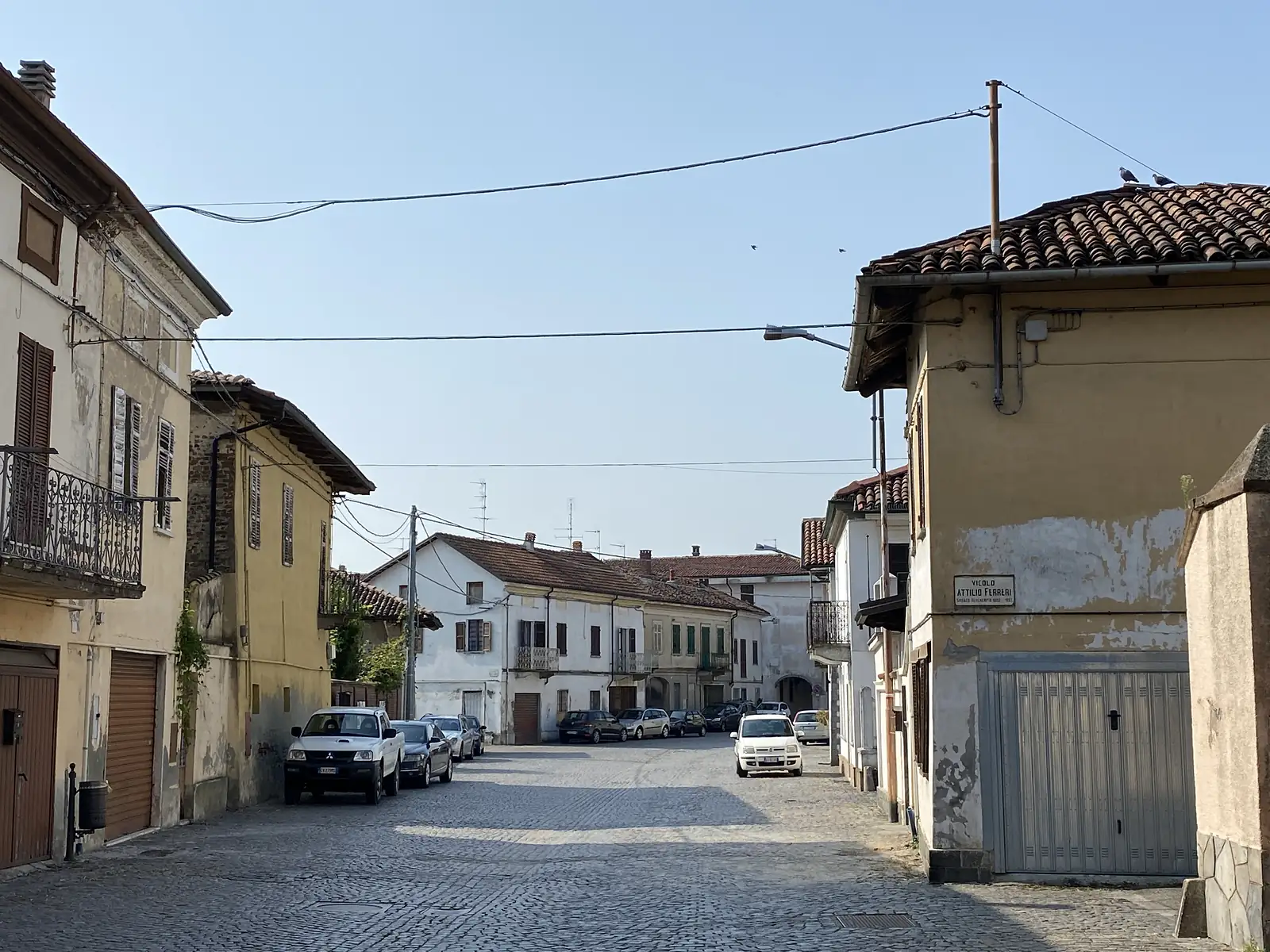 A cobbled village lane lined with old terraced houses and parked cars, marked by the street sign 'Vicolo Attilio Ferreri'.