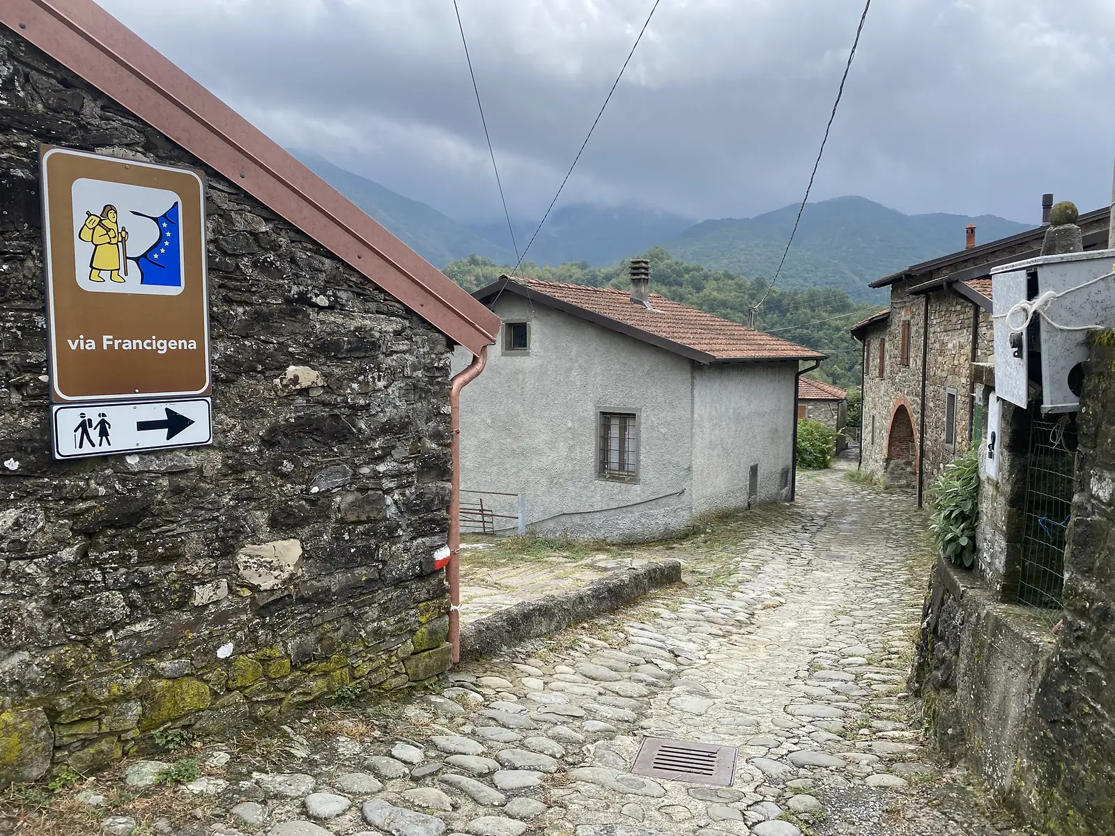 A brown Via Francigena waymark sign, showing a pilgrim figure and an arrow, is fixed to a stone house at the head of a cobbled lane running through a small village.