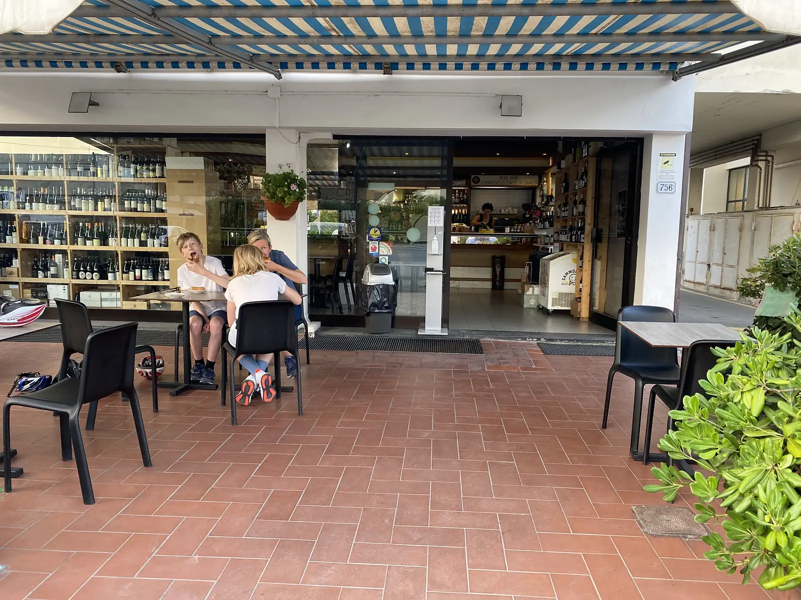 Cyclists eat pastries at an outdoor café table, with cycling helmets resting on a spare chair, in front of a shop window stacked with wine bottles.